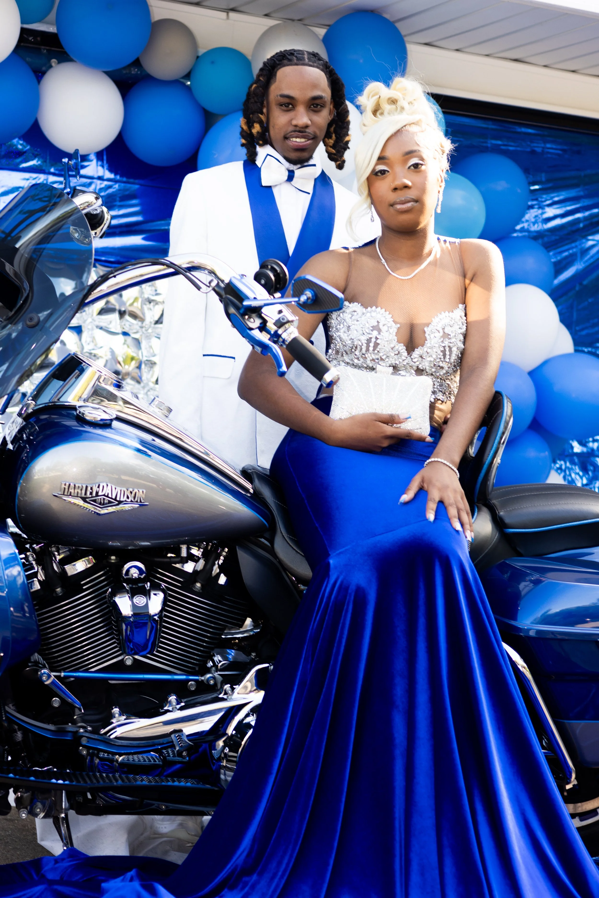 A young woman in a formal blue gown sitting on a Harley Davidson motorcycle, with a young man standing behind her in a white tuxedo, at a celebration backdrop with blue and white balloons.