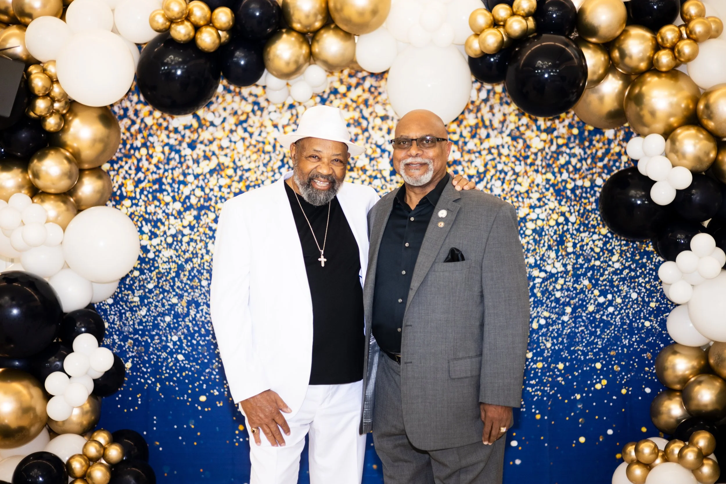 Two men standing in front of a backdrop decorated with balloons in black, white, gold, and multicolored confetti. One man is wearing a white suit and hat, and the other is wearing glasses and a gray suit.