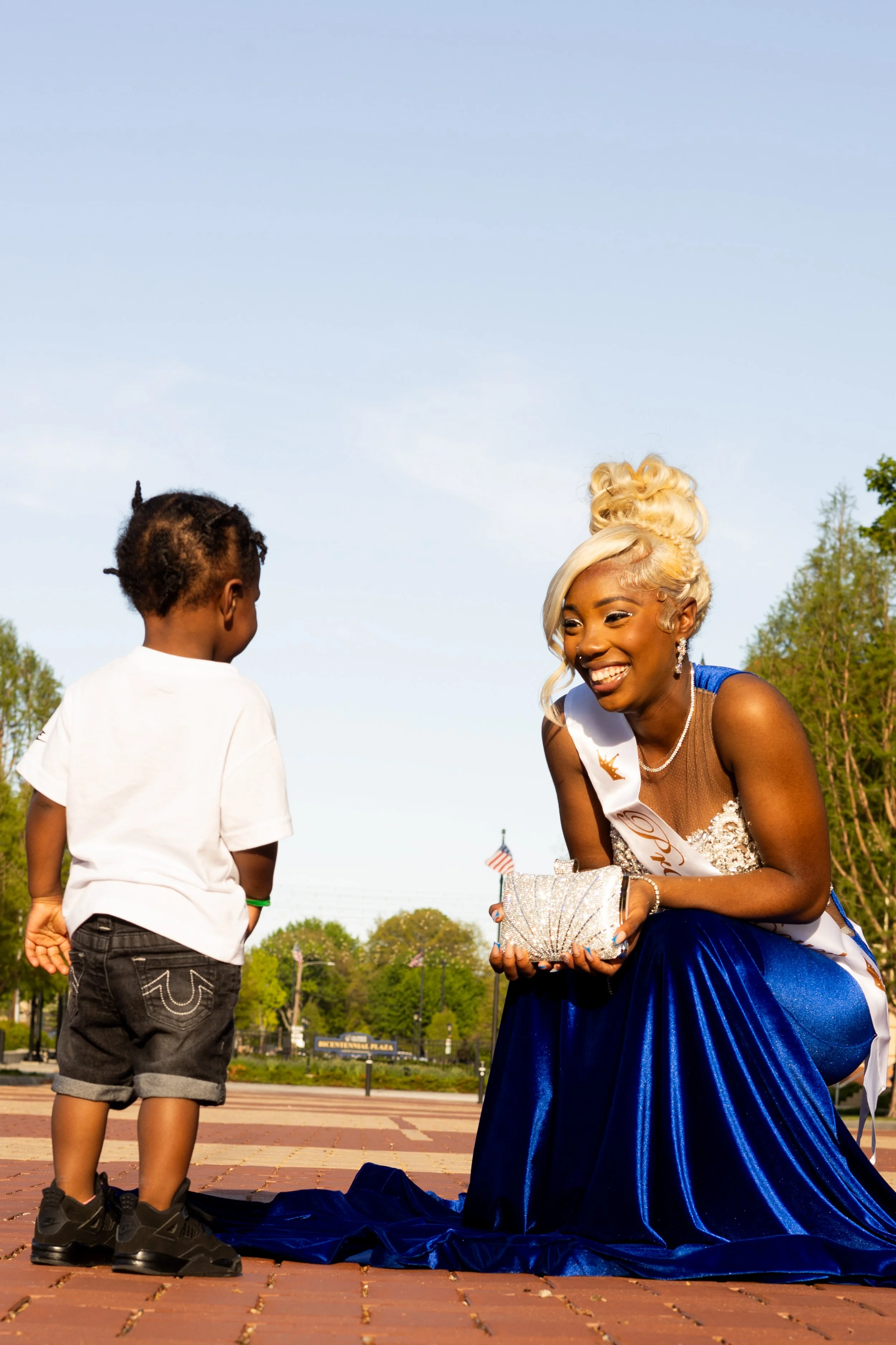 A woman dressed in a royal blue gown and sash, possibly a beauty queen, is smiling and talking to a young boy at an outdoor event. The woman appears to be a pageant winner, holding a sparkling clutch and wearing jewelry, with her hair styled in an elegant updo. The boy has short, curly hair and is wearing a white t-shirt and black shorts. The background shows trees, a blue sky, and an American flag.