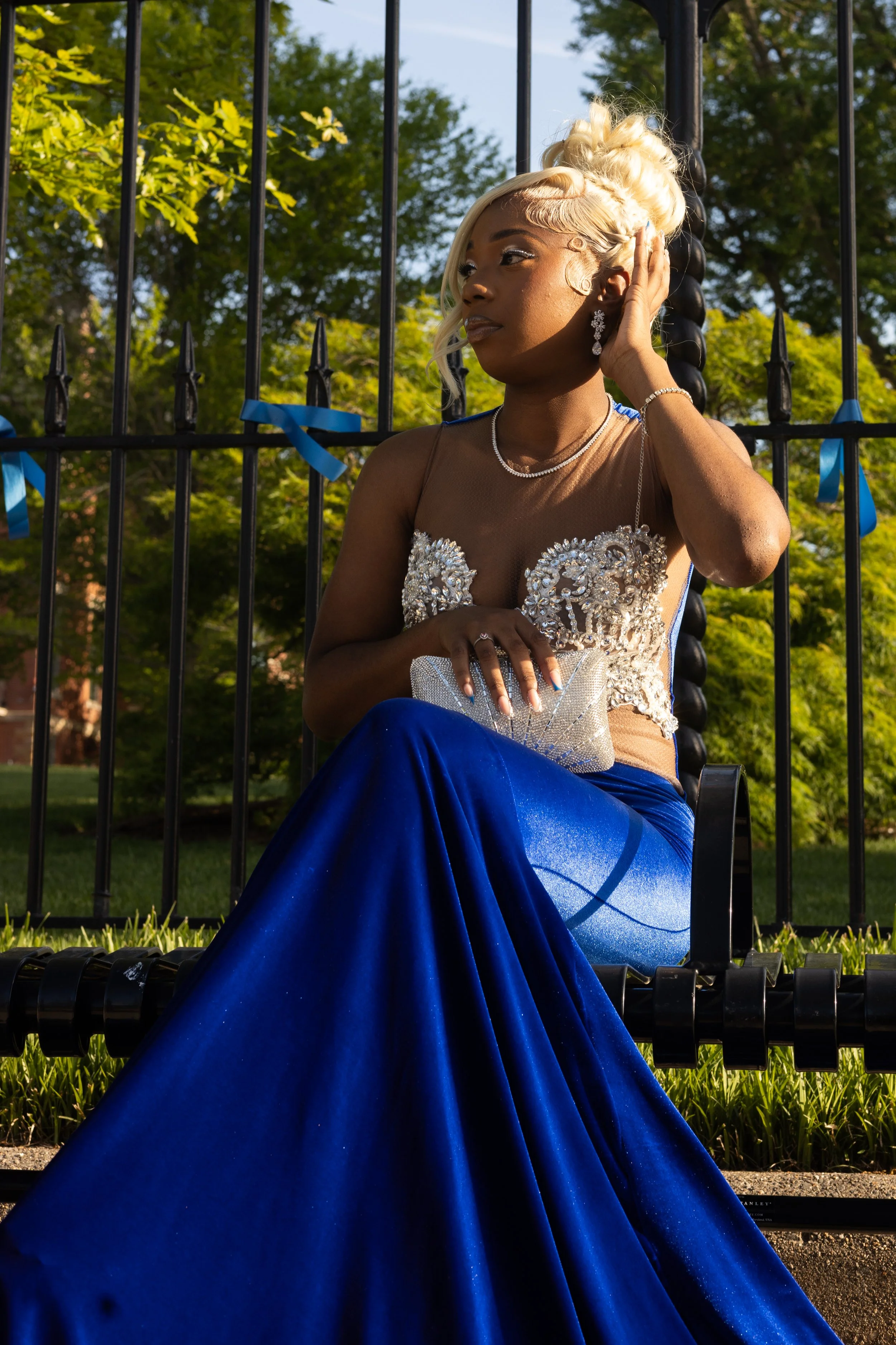 A woman in an elegant dress with intricate beadwork and a blue satin skirt, sitting outdoors near a black wrought-iron fence decorated with blue ribbons, surrounded by green trees under a sunny sky.