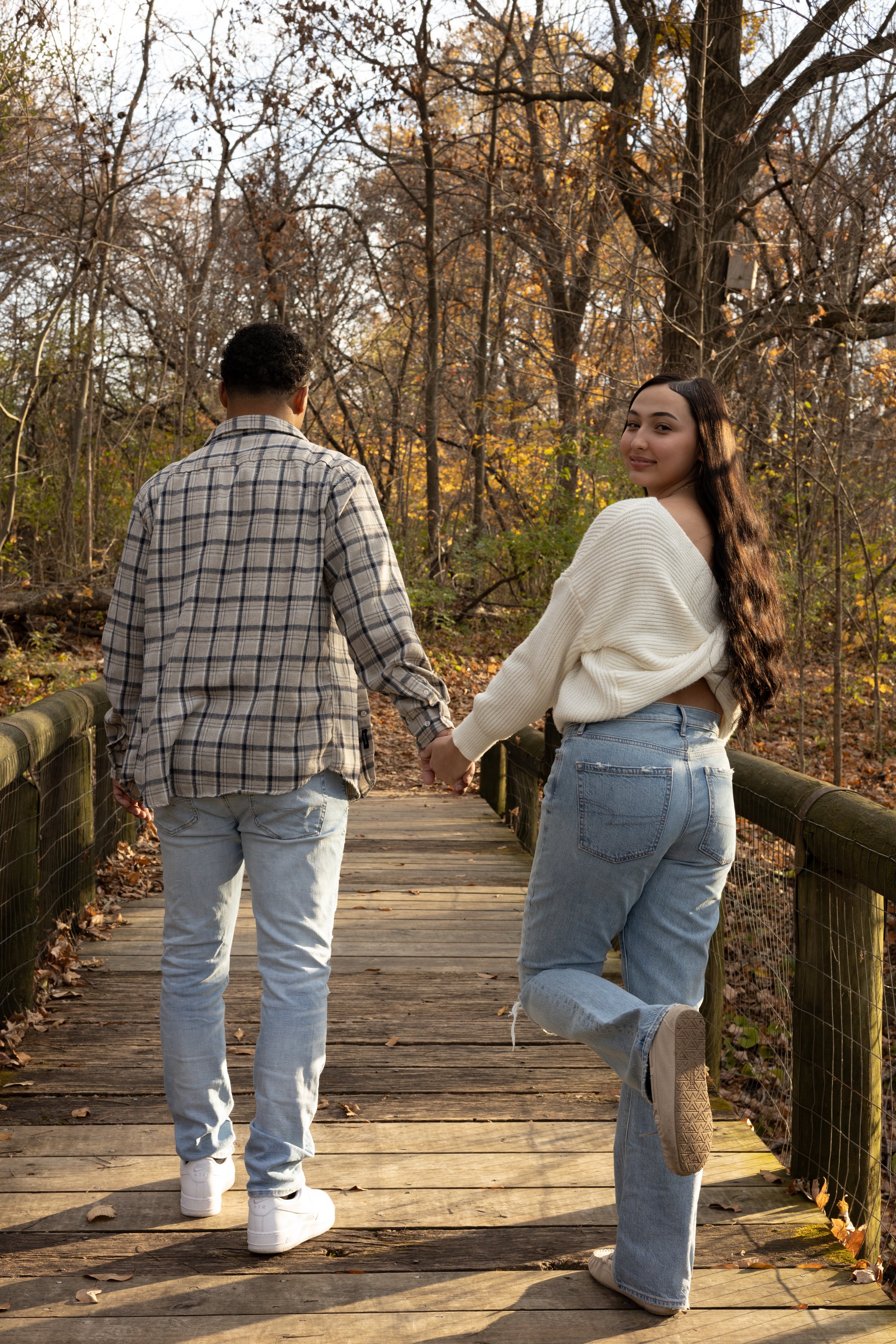 A young woman with long wavy hair and a young man with short curly hair holding hands and walking on a wooden bridge through a forest during autumn.