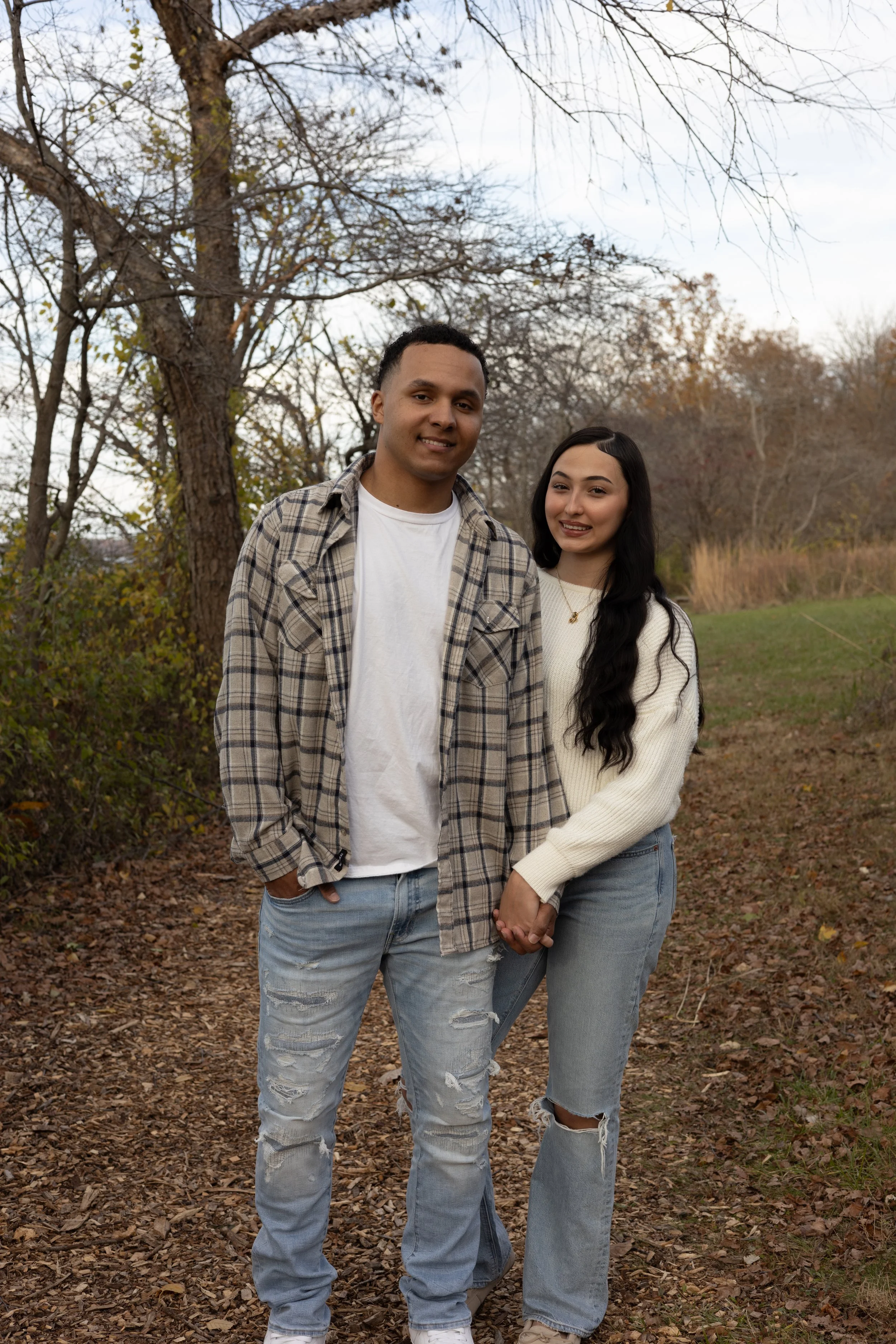 A young man and woman holding hands in an outdoor park during fall, with trees and fallen leaves in the background.