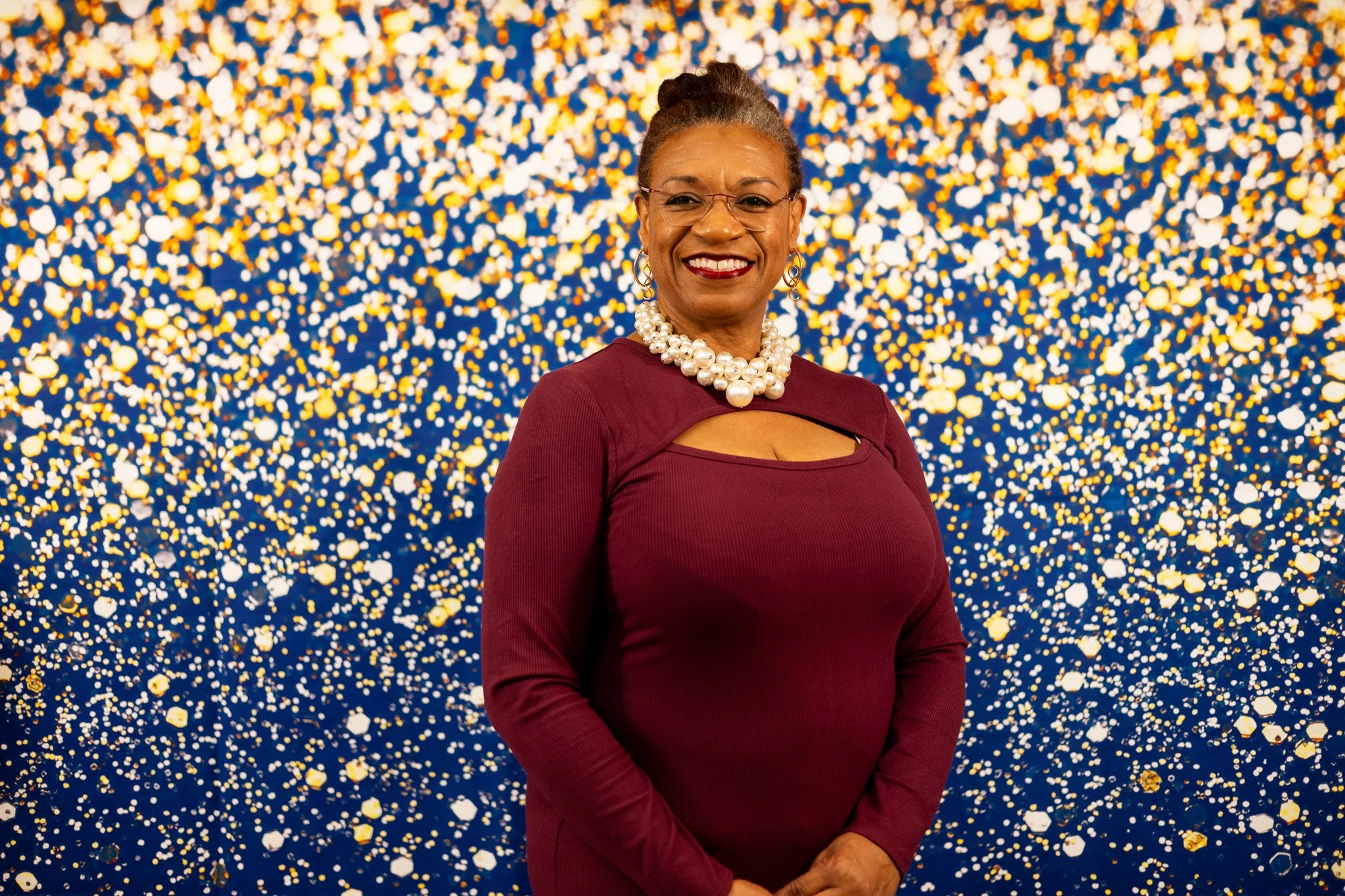 A smiling woman with glasses, pearl necklace, and earrings wearing a maroon dress standing in front of a colorful confetti background.