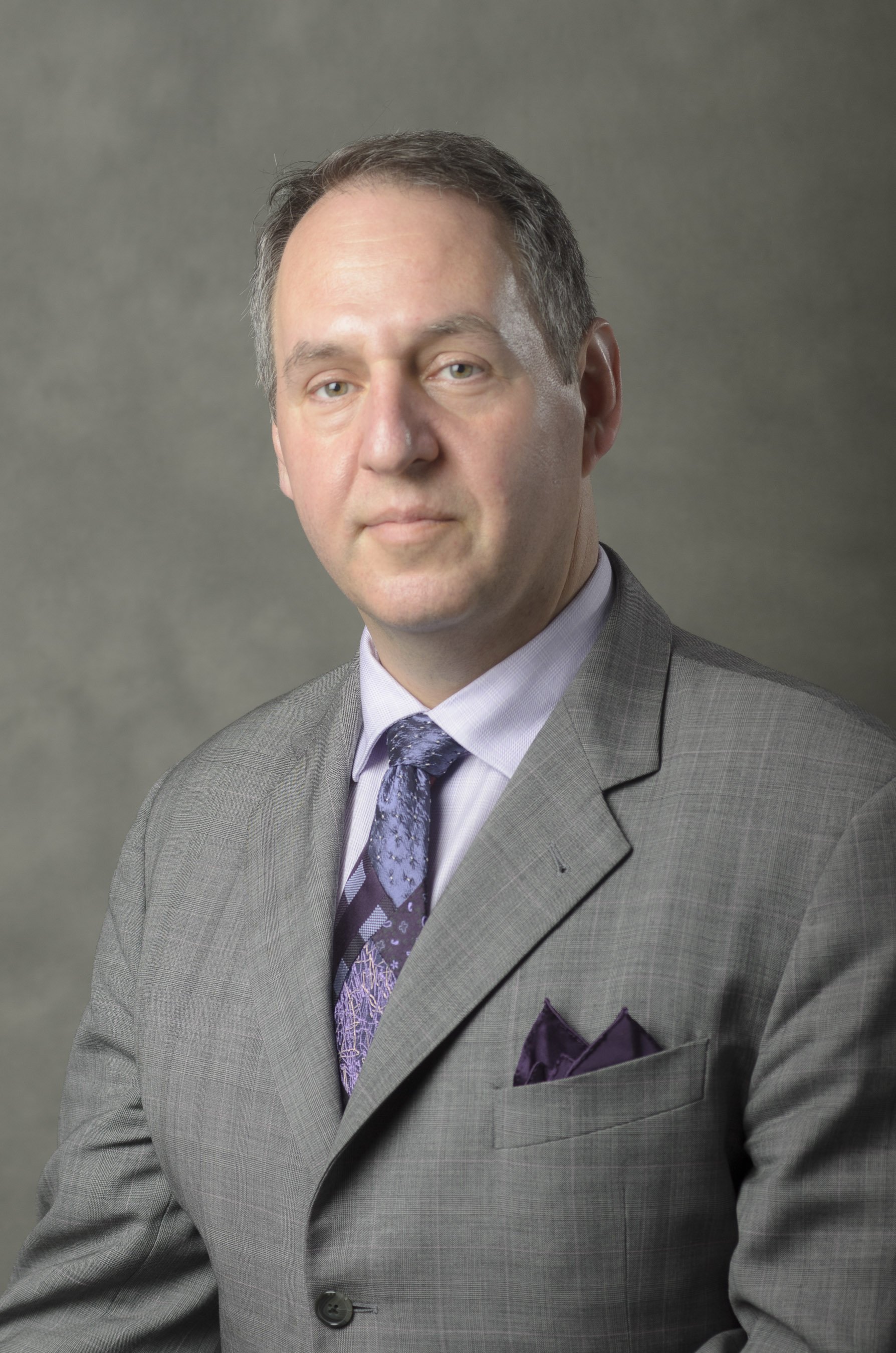 A professional man in a gray suit with a purple patterned tie and pocket square posing against a plain background.