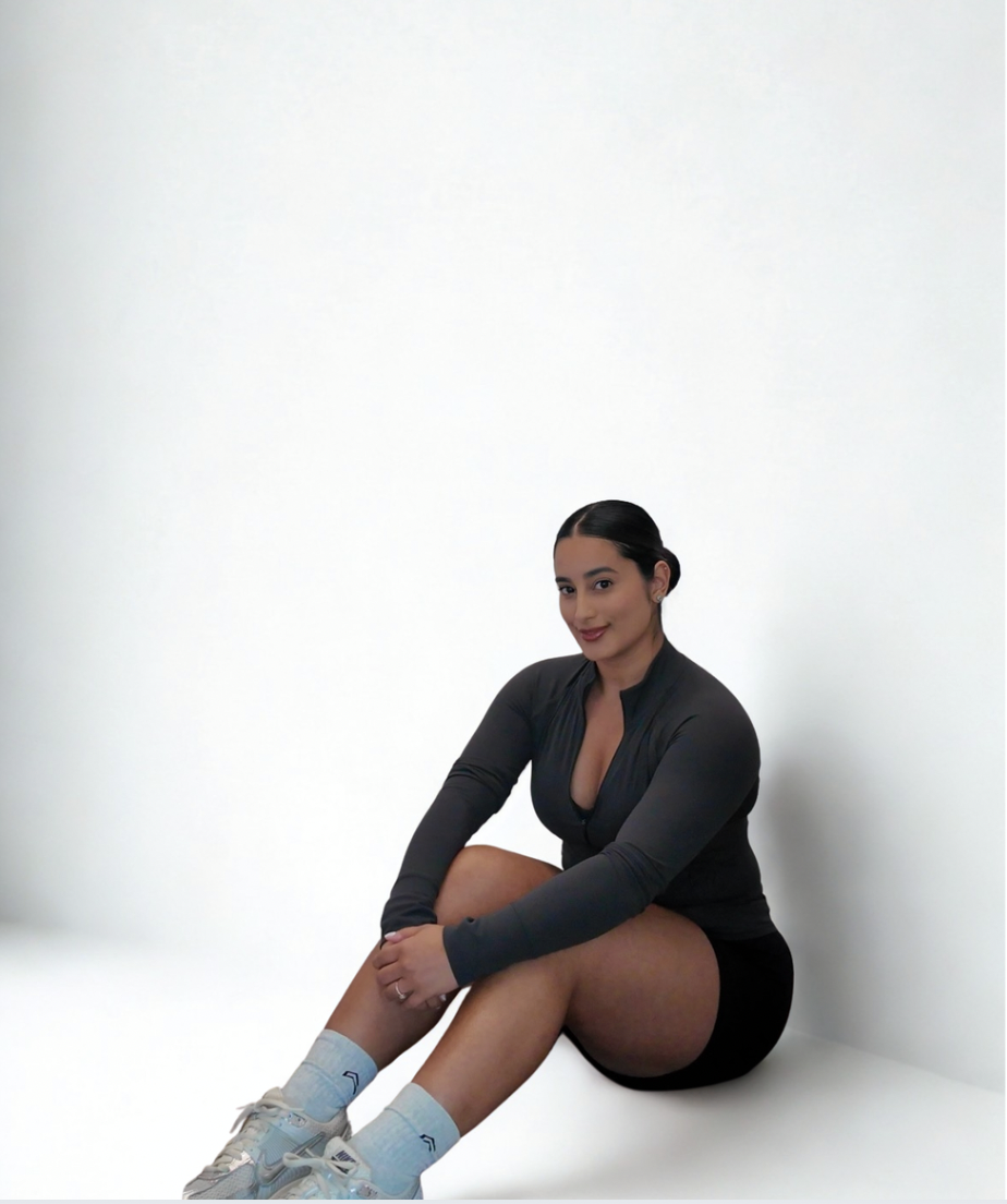 Young woman with dark hair tied back, sitting on the floor with her legs crossed, wearing a black zip-up top, black shorts, white socks, and white sneakers, against a plain white background.