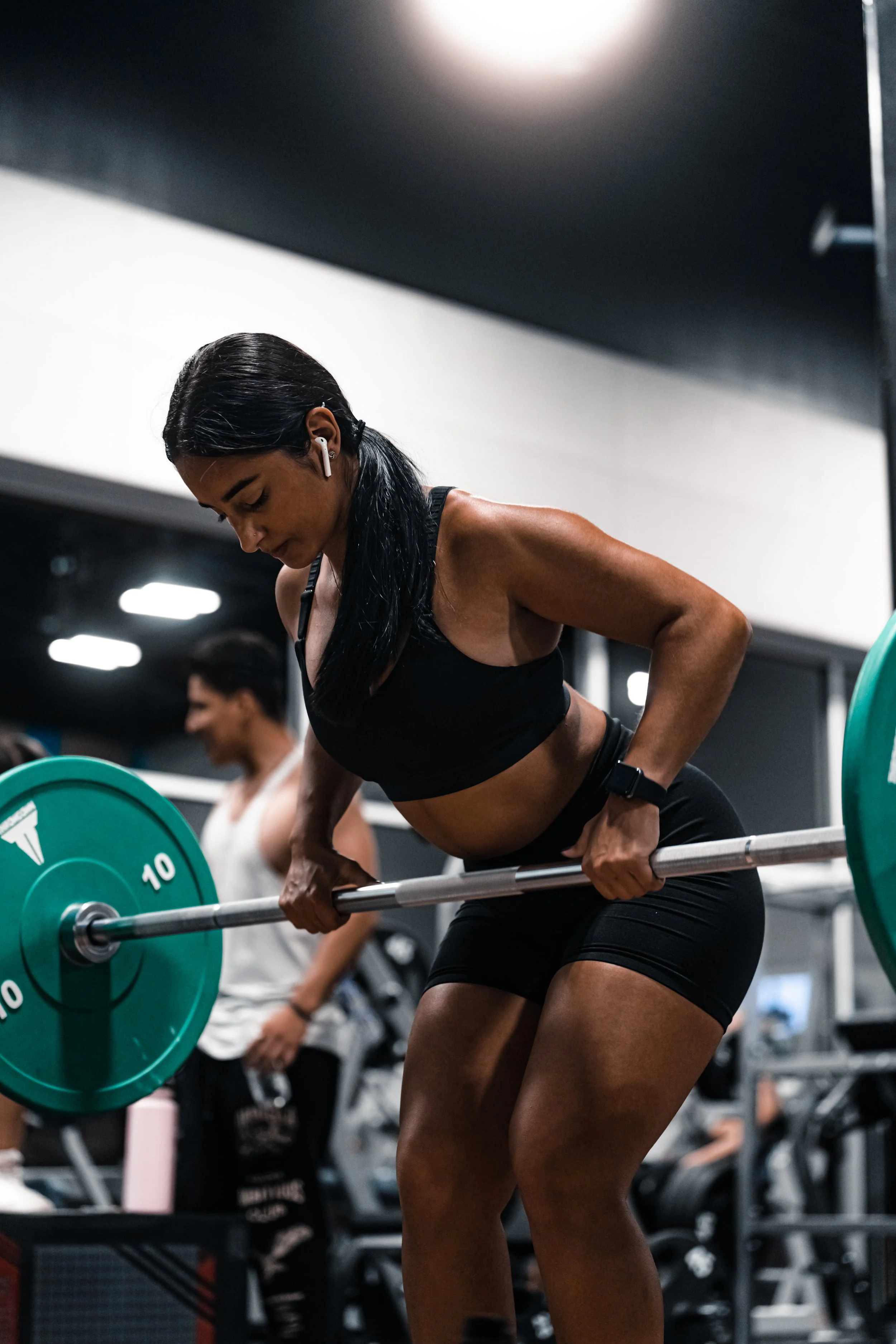 A woman in a gym lifting a barbell with weights, wearing black workout clothes, with headphones, and focused on her exercise.