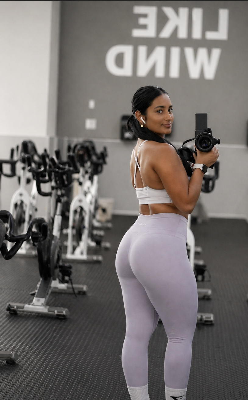 A woman taking a selfie in a gym, wearing a white sports bra and leggings, with gym bikes in the background and a motivational sign on the wall.