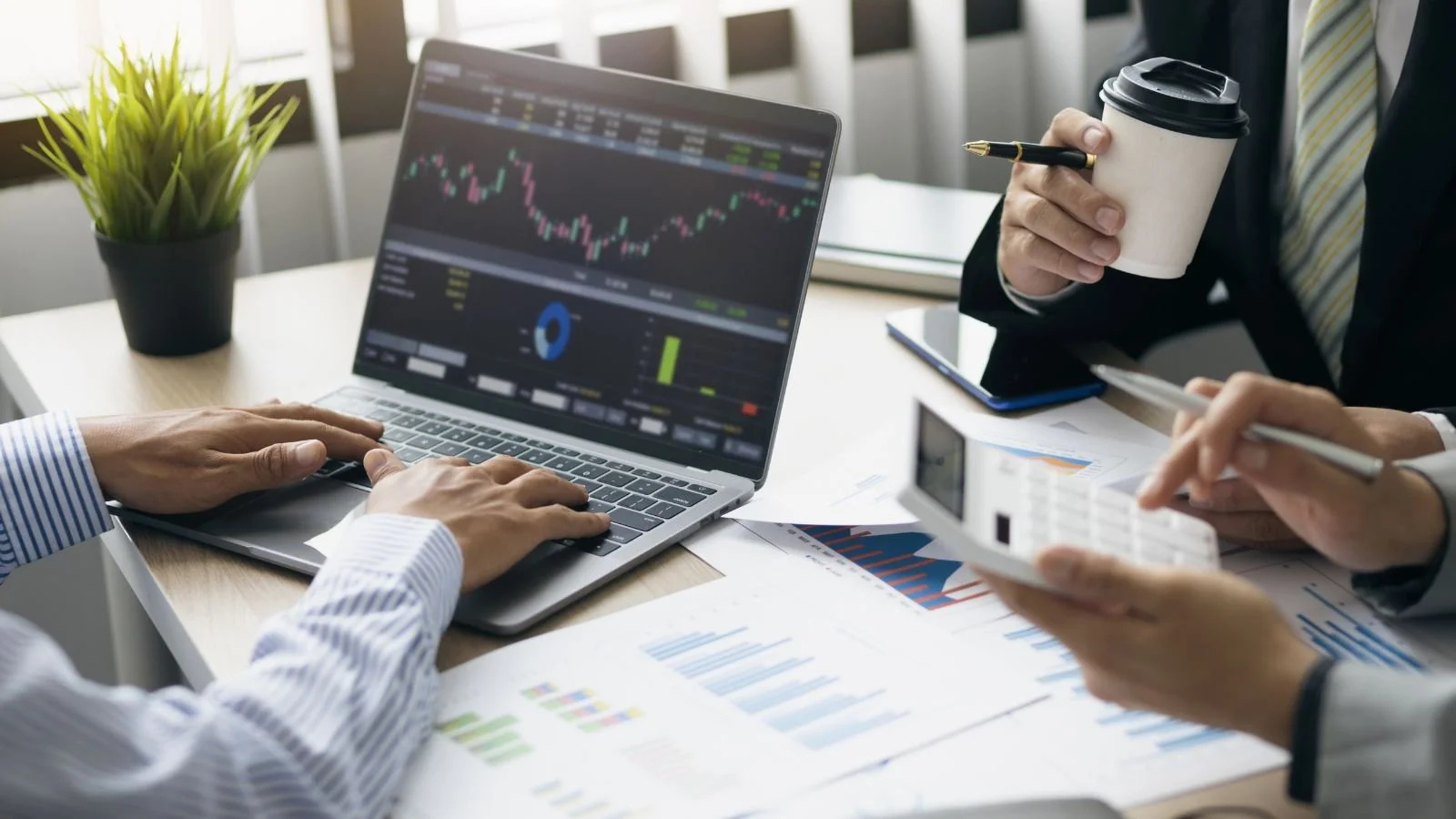 Businesspeople analyzing financial data on charts and graphs at a meeting, with a laptop displaying stock market trends and a person holding a coffee cup.