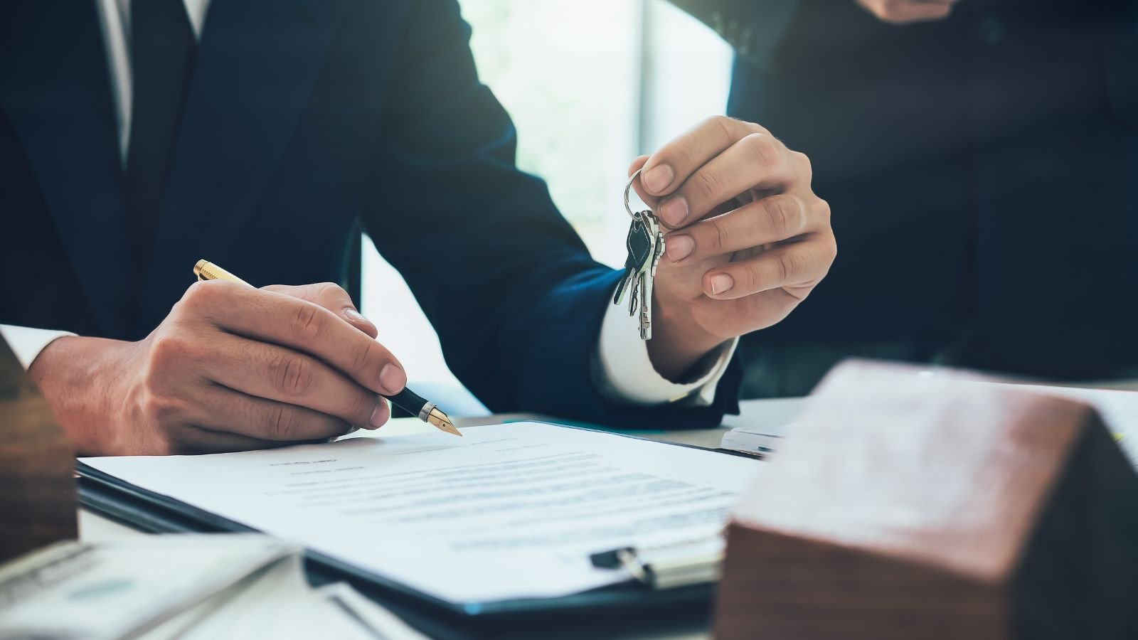 Two individuals in suits exchanging and signing legal documents, with one person holding a set of keys.