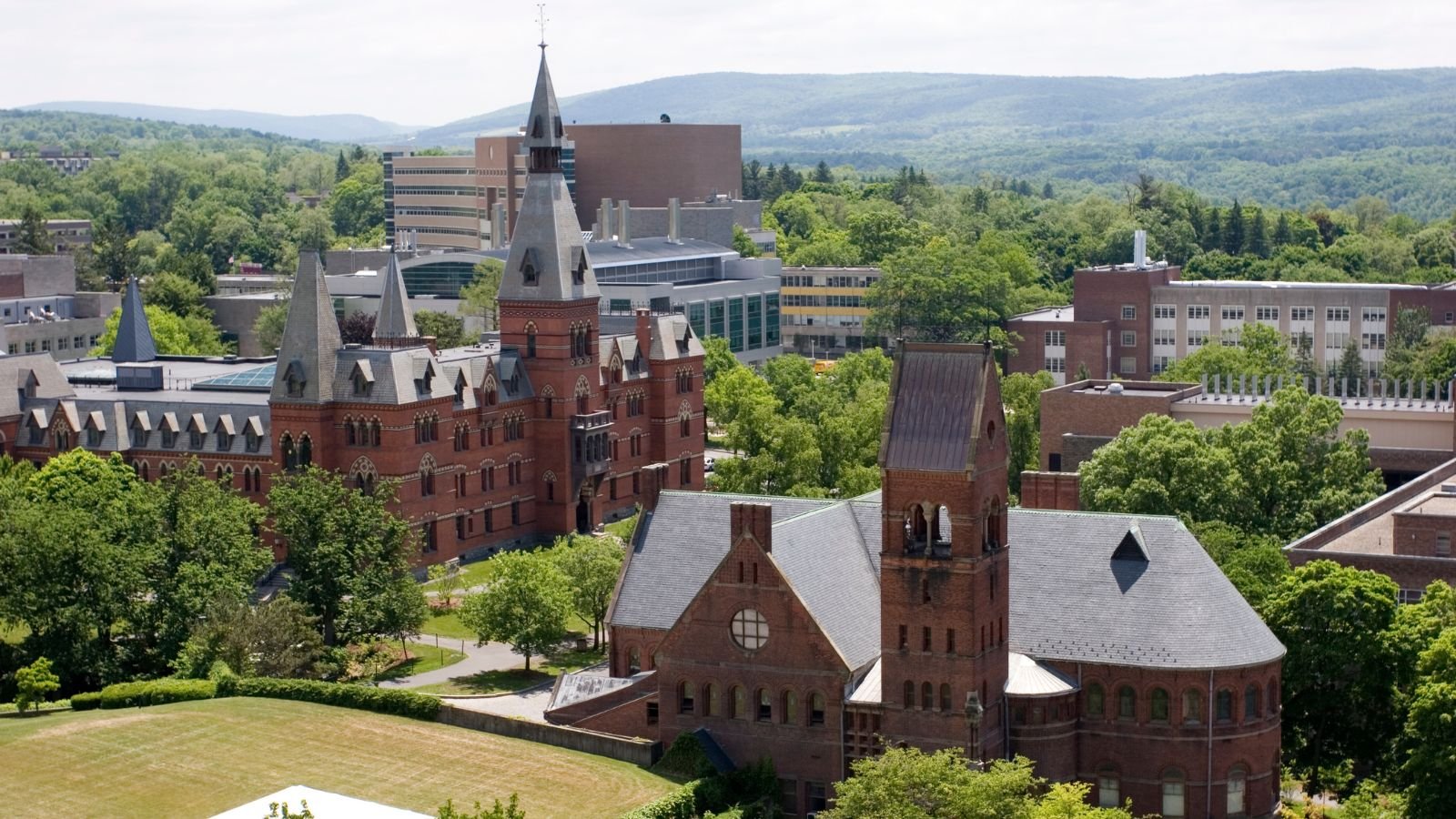 Aerial view of a historic brick church with multiple steeples, surrounded by green trees, in a cityscape with modern buildings and distant hills.