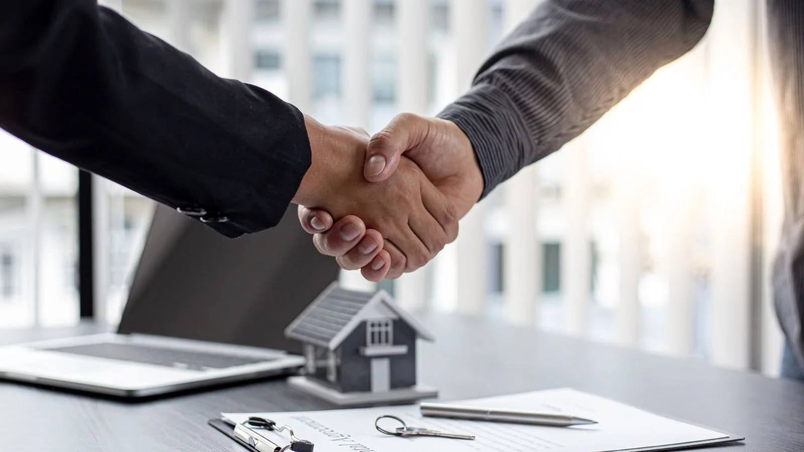 Two people shaking hands over a table with a small house model, a clipboard, keys, a pen, and a tablet in a bright office setting.