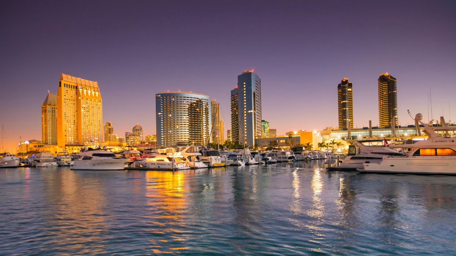 City skyline with high-rise buildings and yachts docked at a marina during sunset.