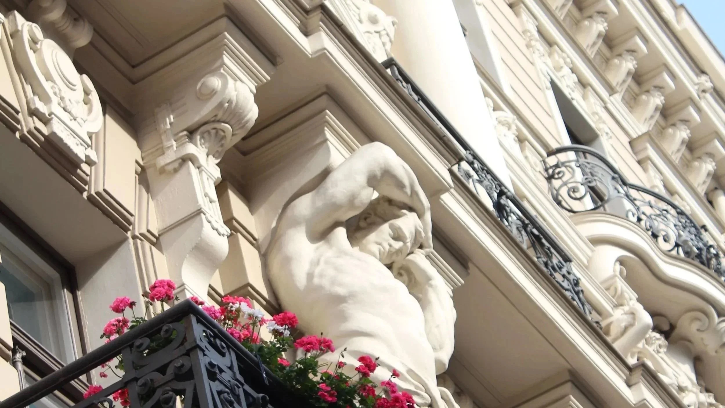 Close-up of an ornate building facade with detailed architectural elements, including sculptures and iron balcony railing, with pink flowers on the balcony.
