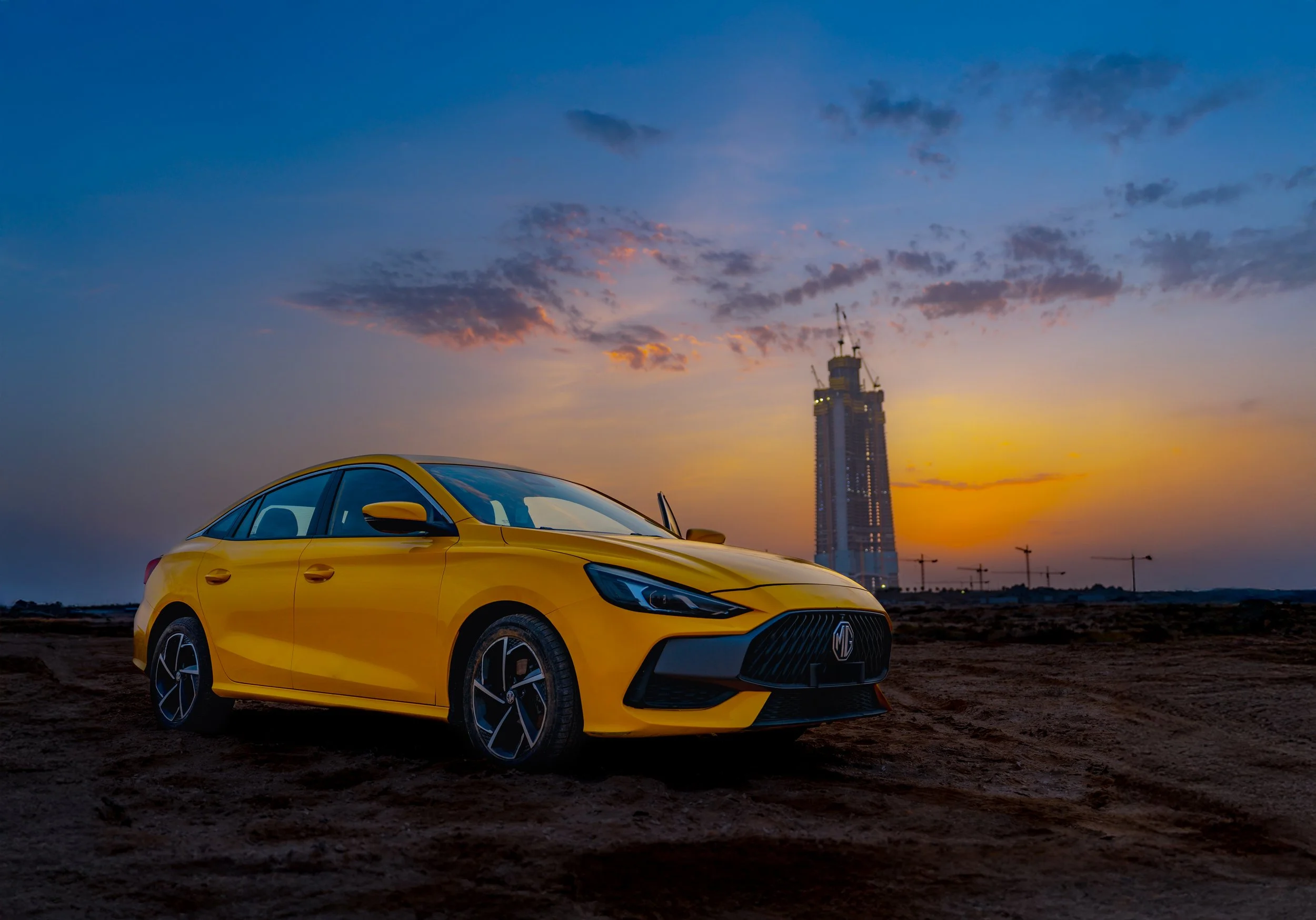 A yellow sports car parked on a dirt surface during sunset, with a city skyline and skyscraper in the background, and a partly cloudy sky with shades of blue, orange, and pink.