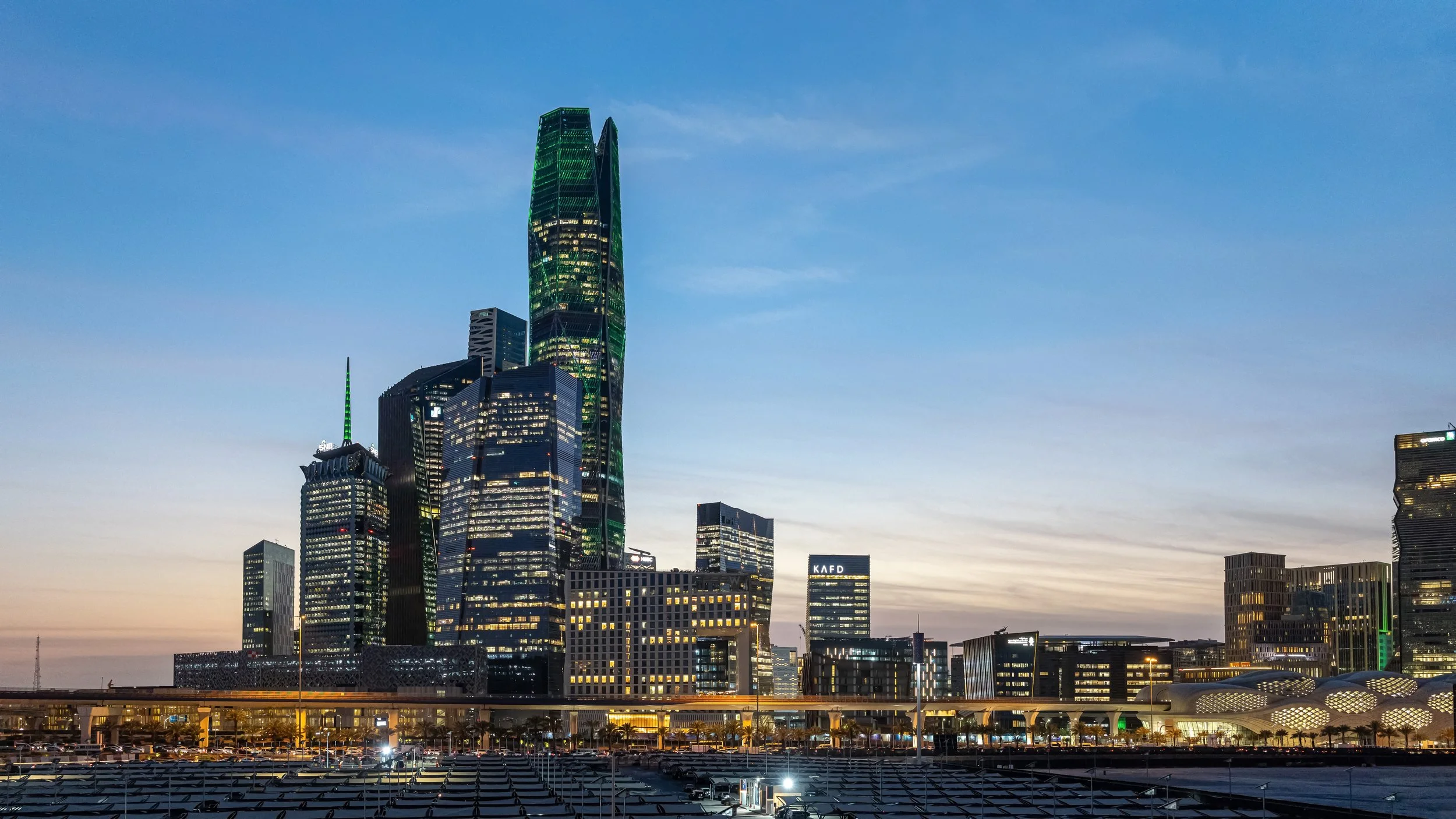 Skyline of a modern city during dusk with tall skyscrapers, including one with green lighting accents, and a parking lot in the foreground.