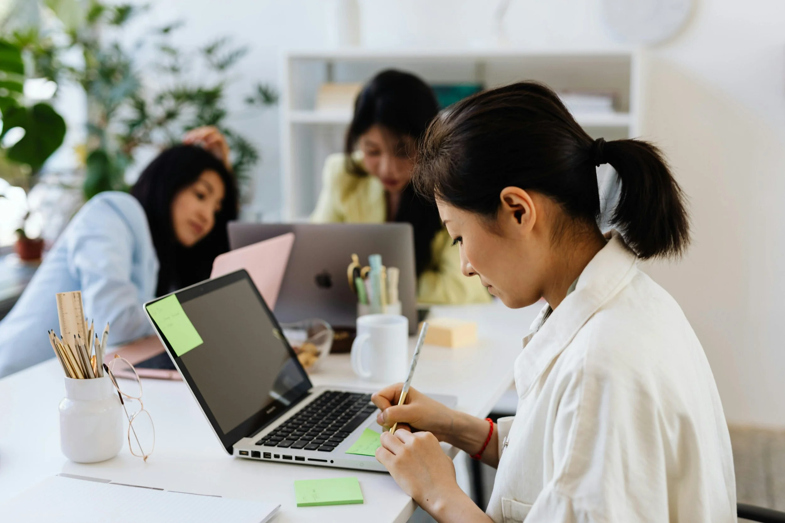 Three women working at a desk with laptops, sticky notes, and stationery, in a bright office with plants and shelves in the background.