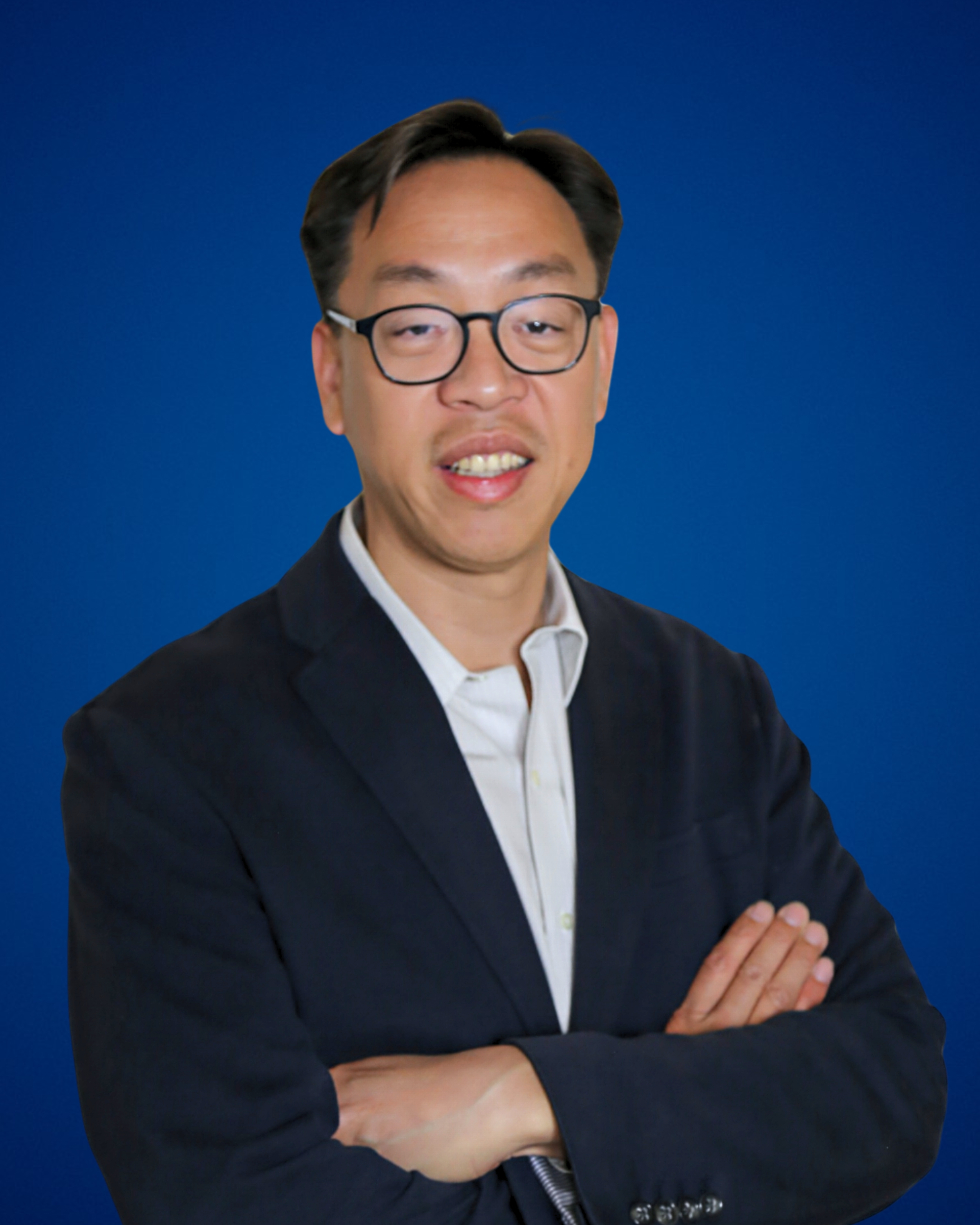 A man wearing glasses and a dark suit with a white shirt, standing with arms crossed against a solid blue background.