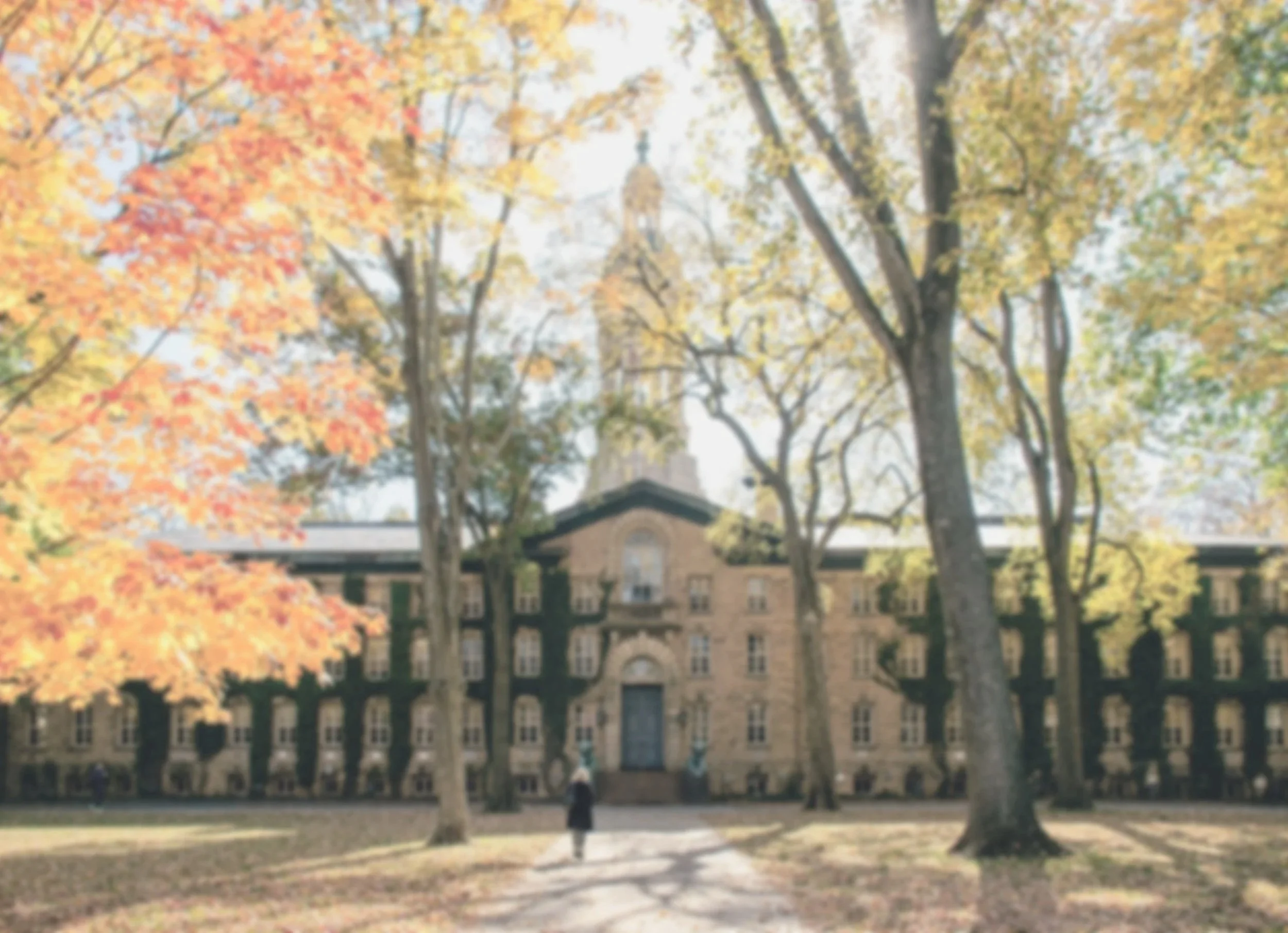 A person walking towards a large brick building with multiple windows, surrounded by trees with yellow and orange autumn leaves.