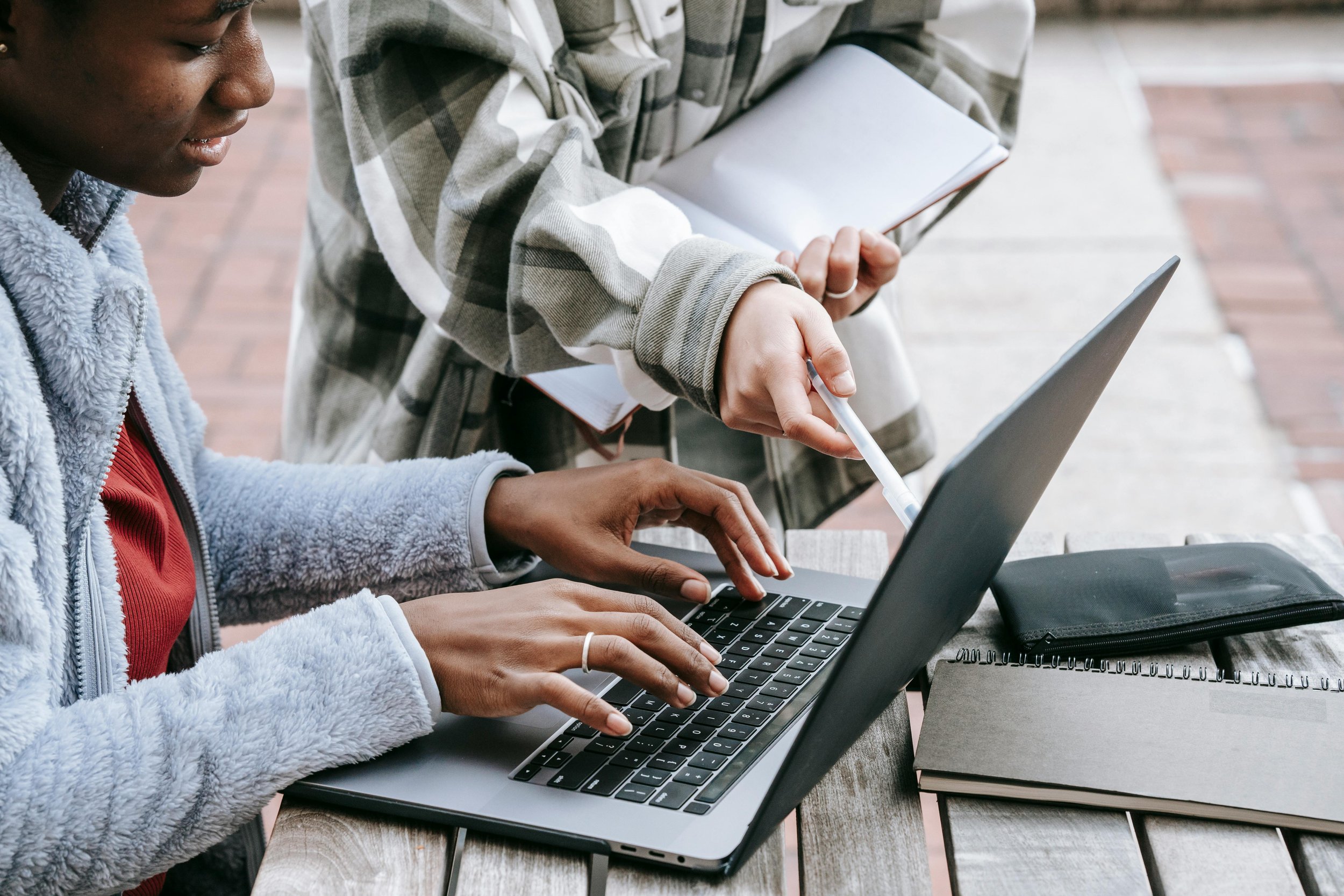 Two people collaborating outdoors, one using a laptop and the other using a tablet, with notebooks nearby.