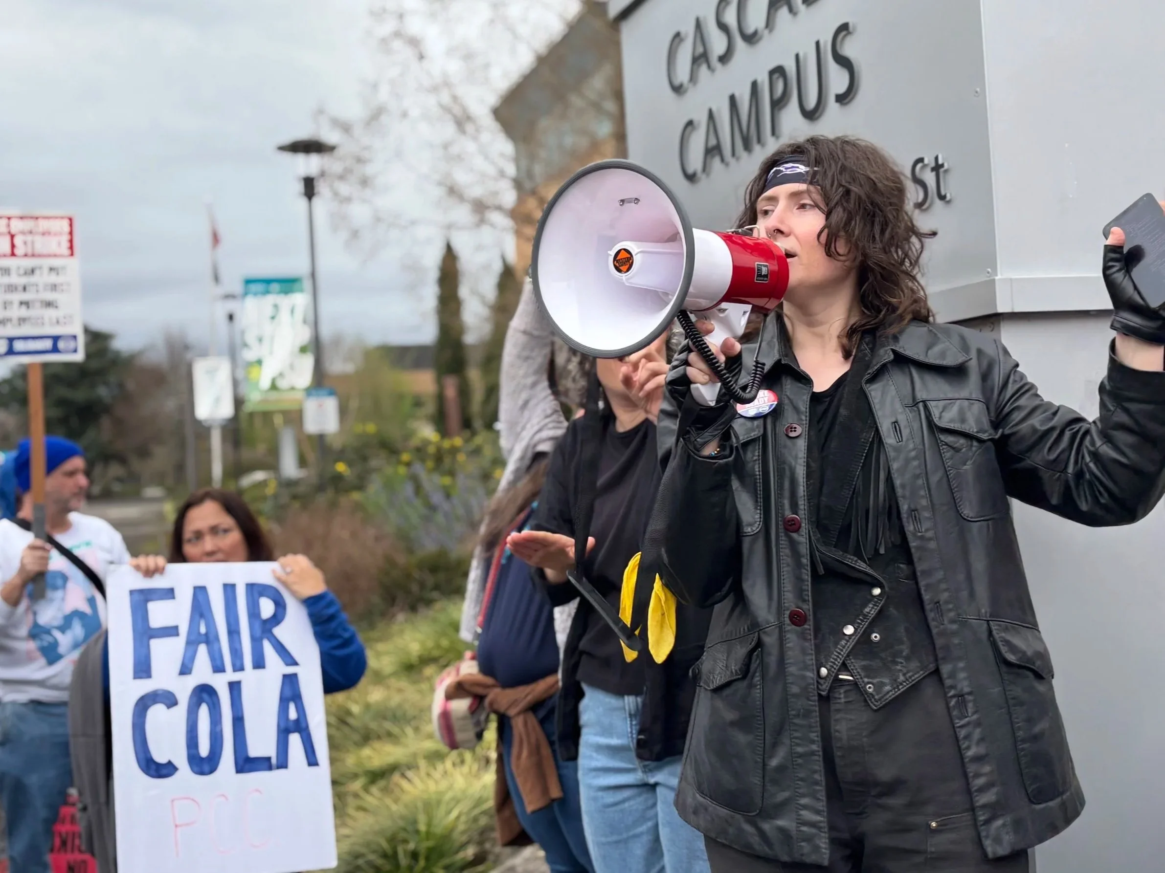 Photo of Aenghus using a megaphone and  leading a huddle at the Cascade Campus picket line. Aenghus is dressed in a black leather vest and headband for 80s theme day.