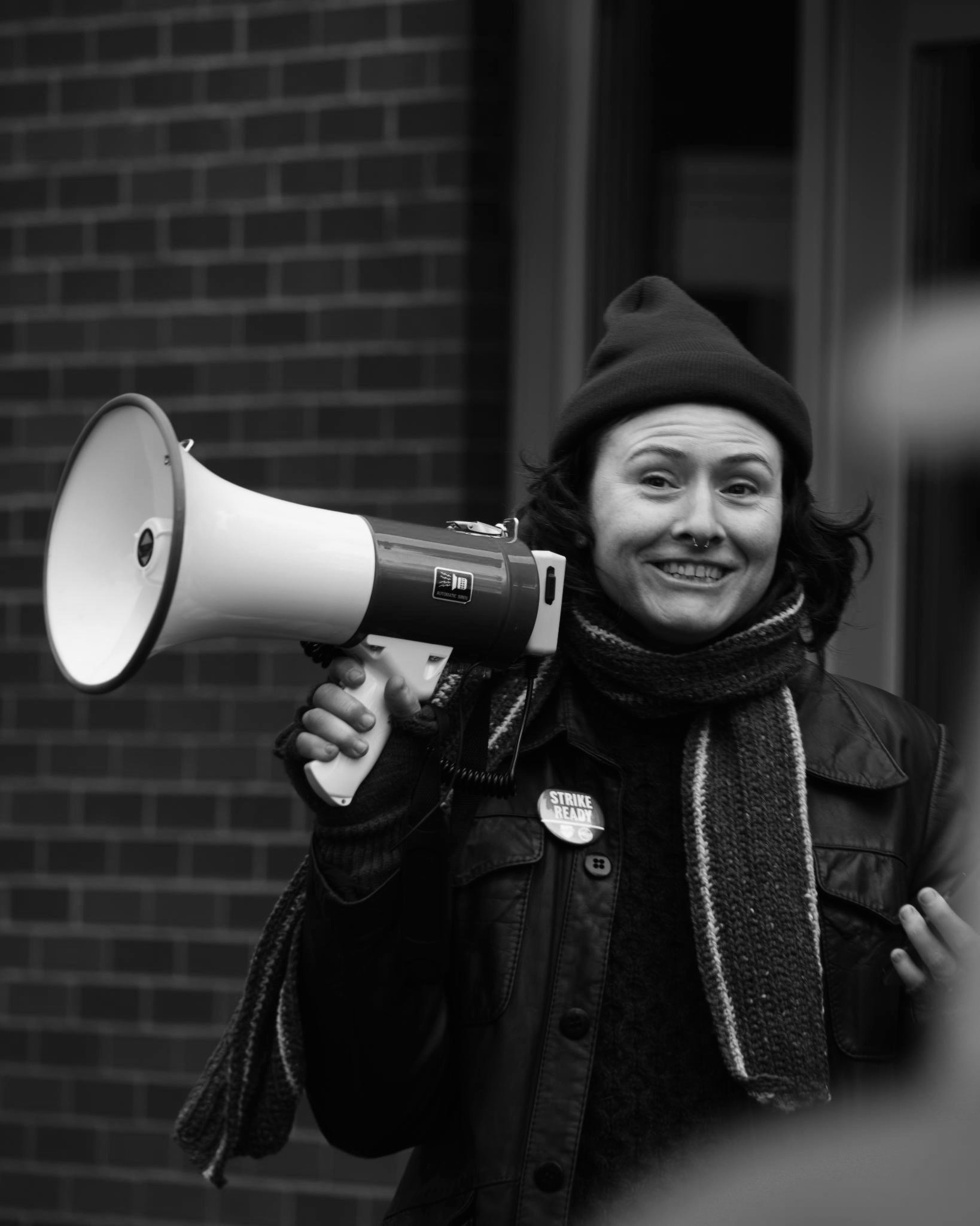 Black and white photo of Aenghus in a hat and scarf, carrying a megaphone at a picket line.