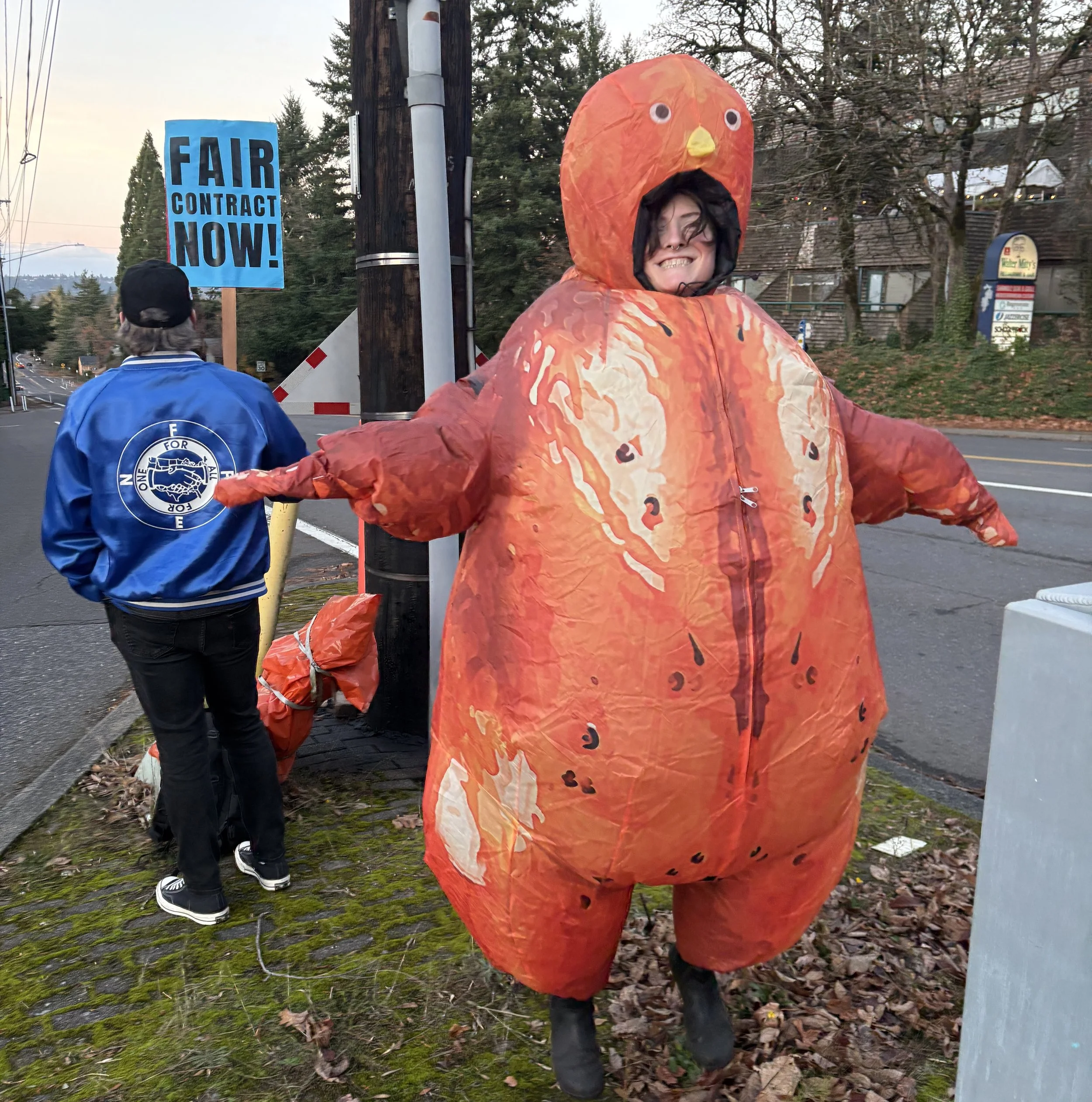 Photo of Aenghus in a blow-up chicken costume at the 2026 SY Practice Picket. Person behind Aenghus is carrying a sign that says "Fair Contract Now!"