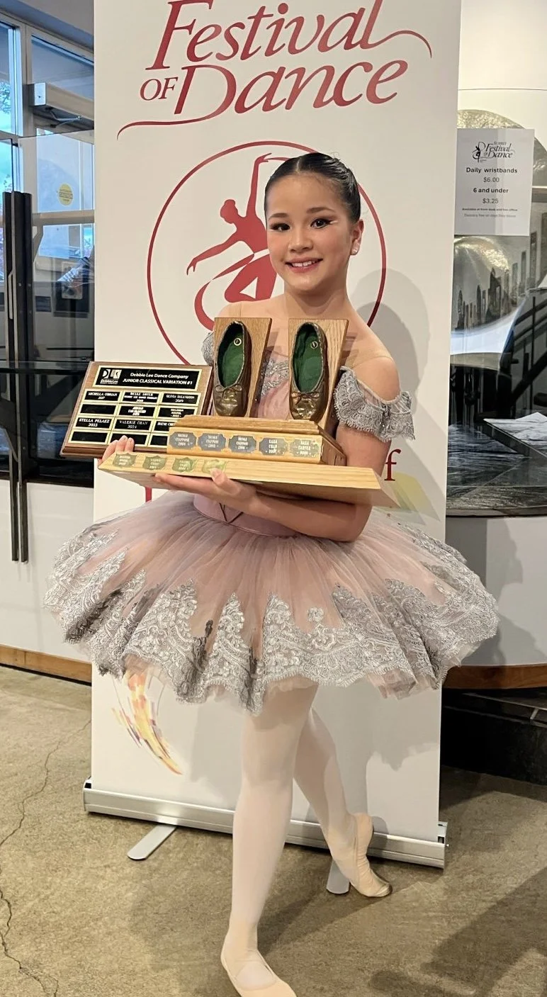 A young girl dressed as a ballet dancer holding a wooden box with a pair of small ballet shoes and a plaque, standing in front of a sign for the Festival of Dance.