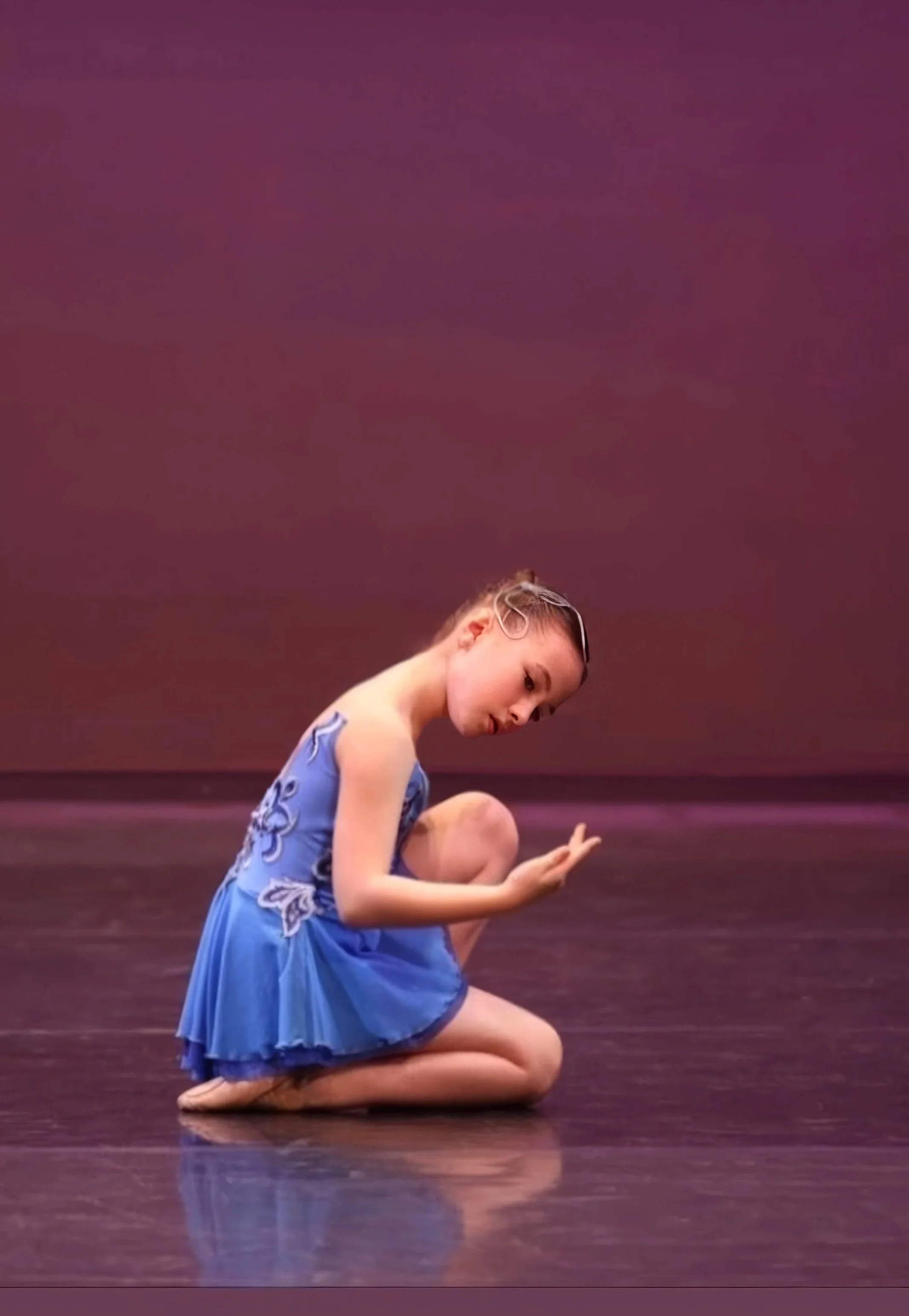 A young girl wearing a blue dress kneeling on a stage, with a purple background, gazing downward with a focused expression.