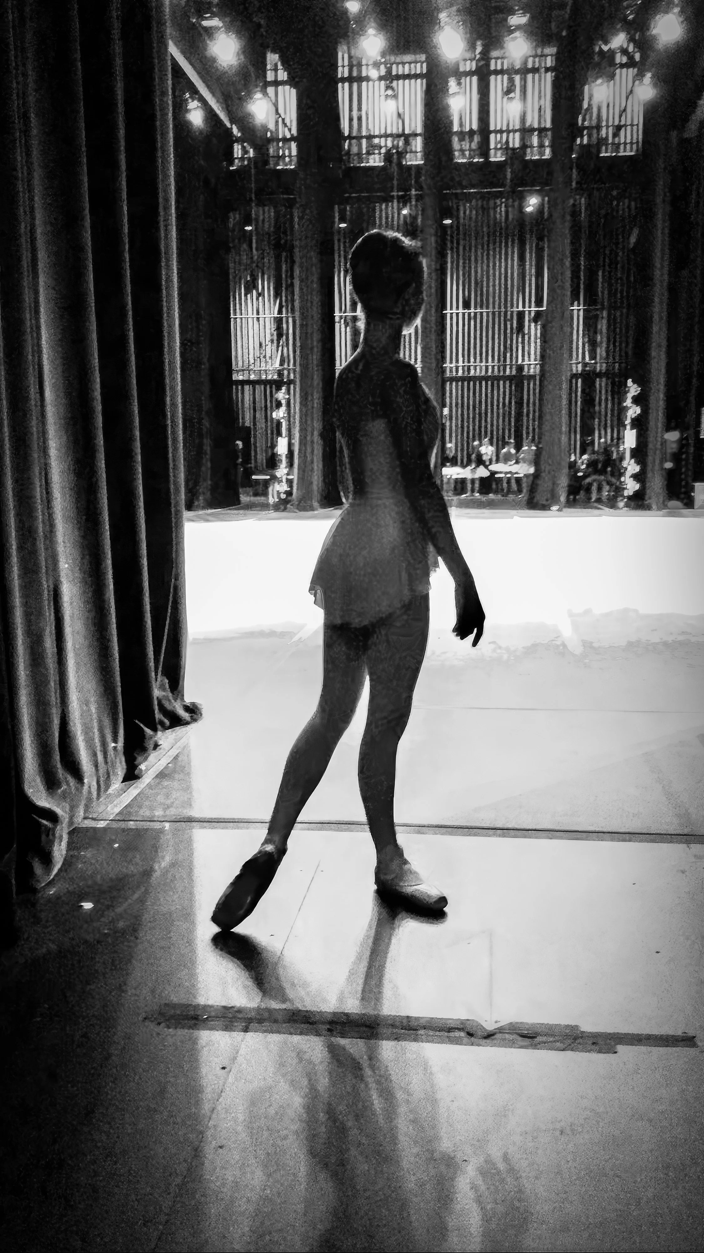 A silhouette of a ballet dancer standing on stage, viewed from behind, with a backdrop of vertical bars and stage lighting.