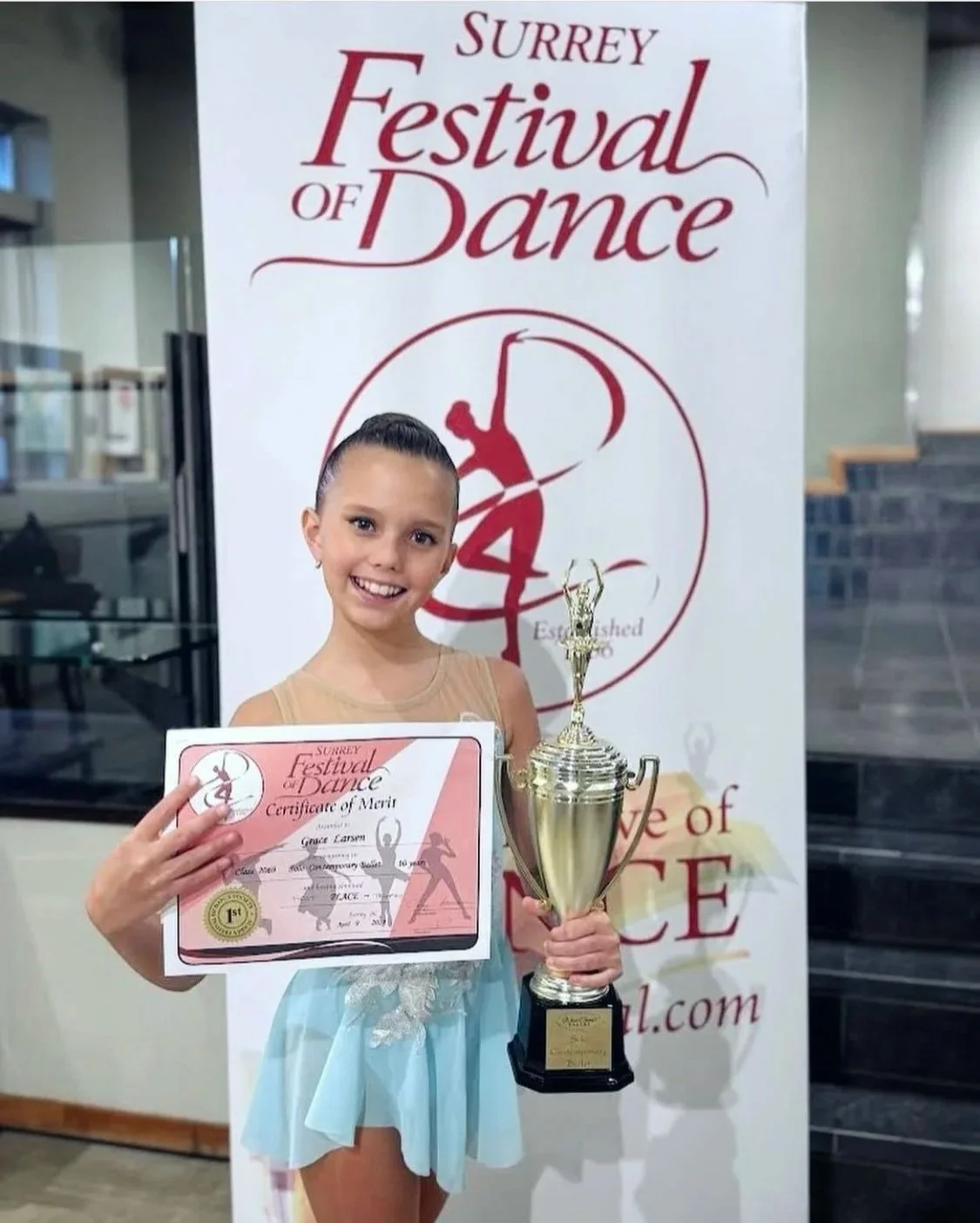 A young girl in a light blue dance costume holding a trophy in one hand and a certificate of merit in the other, smiling, at the Surrey Festival of Dance event.