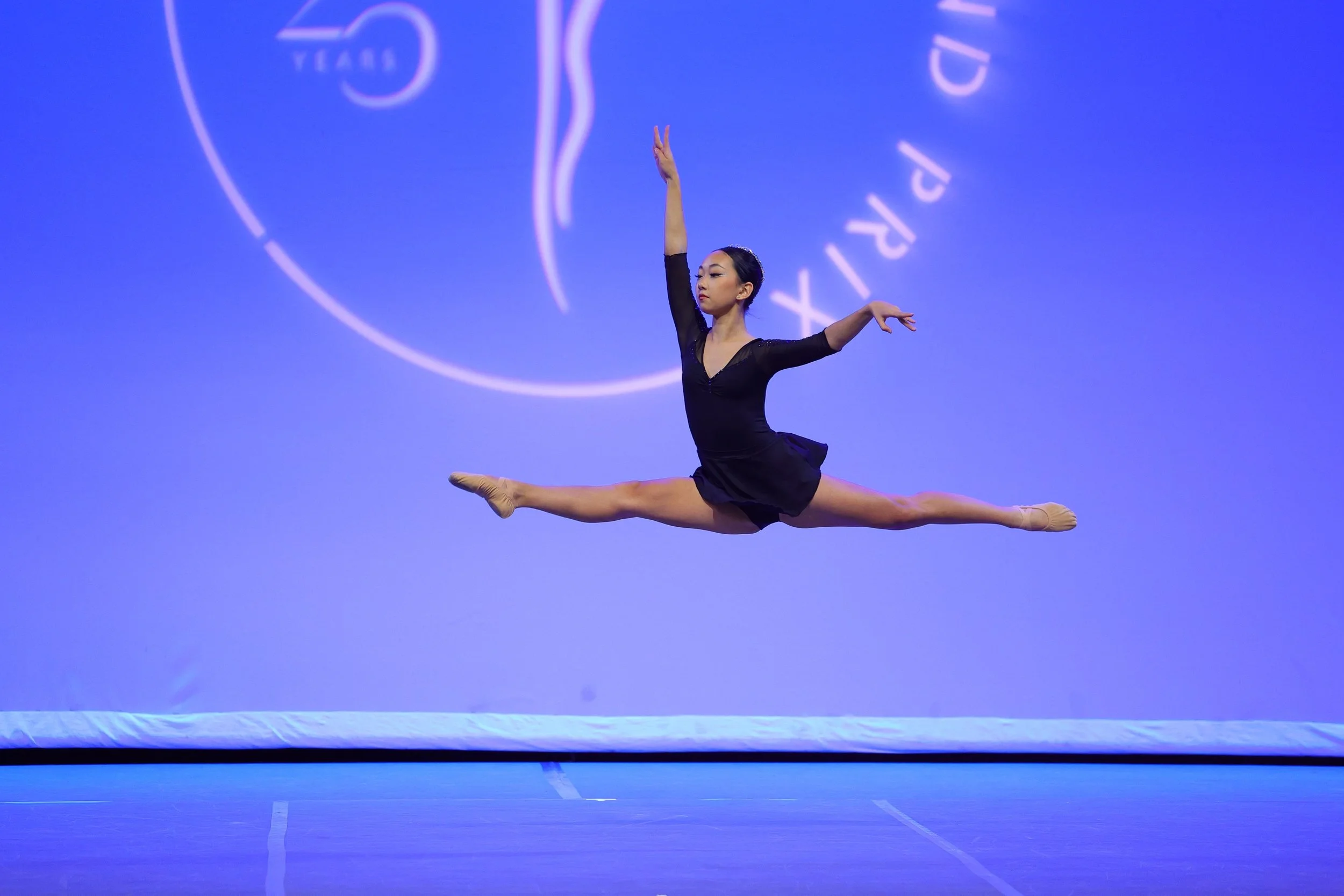 A female gymnast performing a split leap in mid-air during a routine on a blue stage with a large logo projected in the background.
