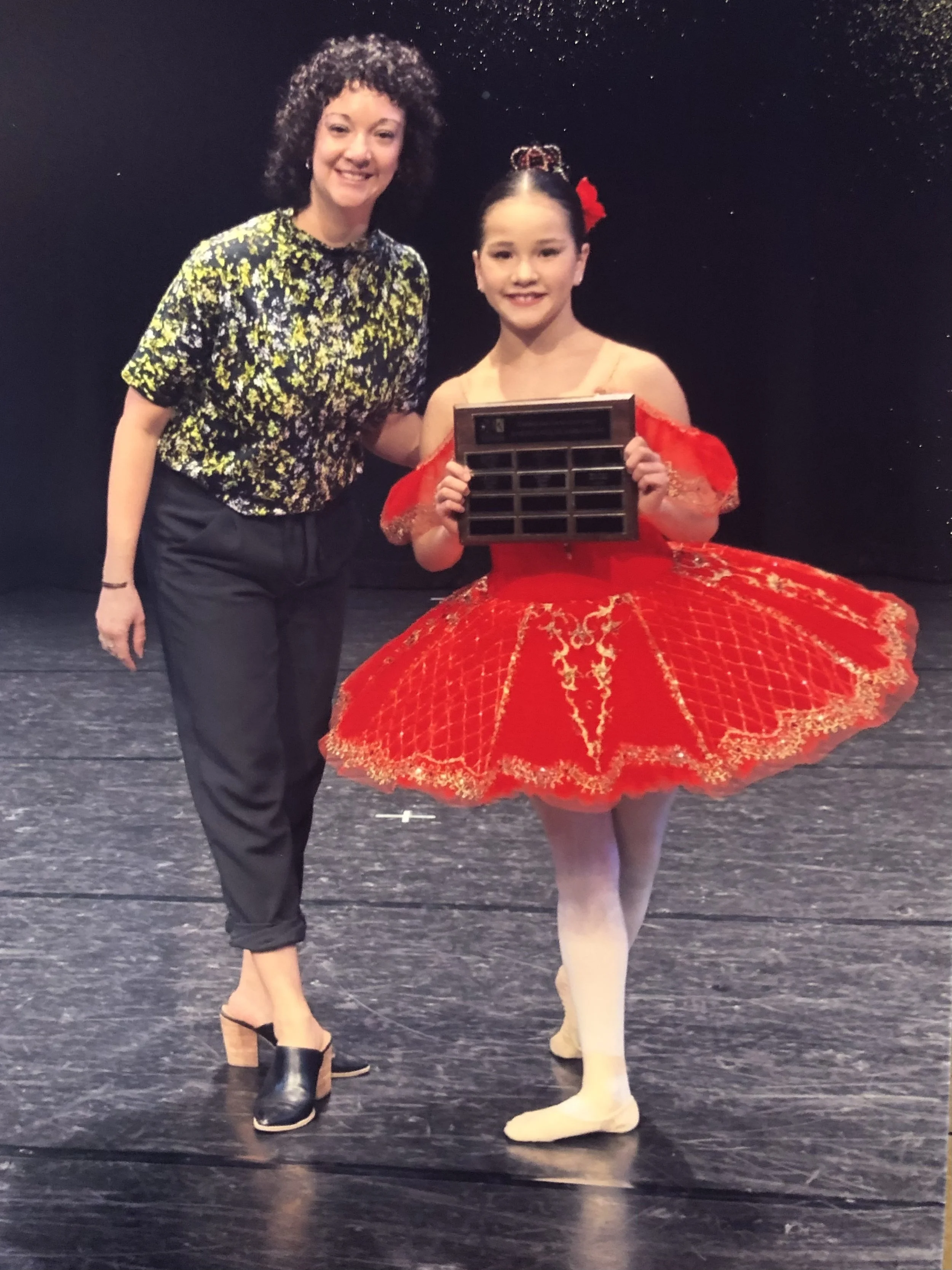A young girl dressed in a red ballet costume with a large, decorated tutu, standing on stage with another woman. The girl is holding a wooden award or plaque, and both are smiling at the camera.