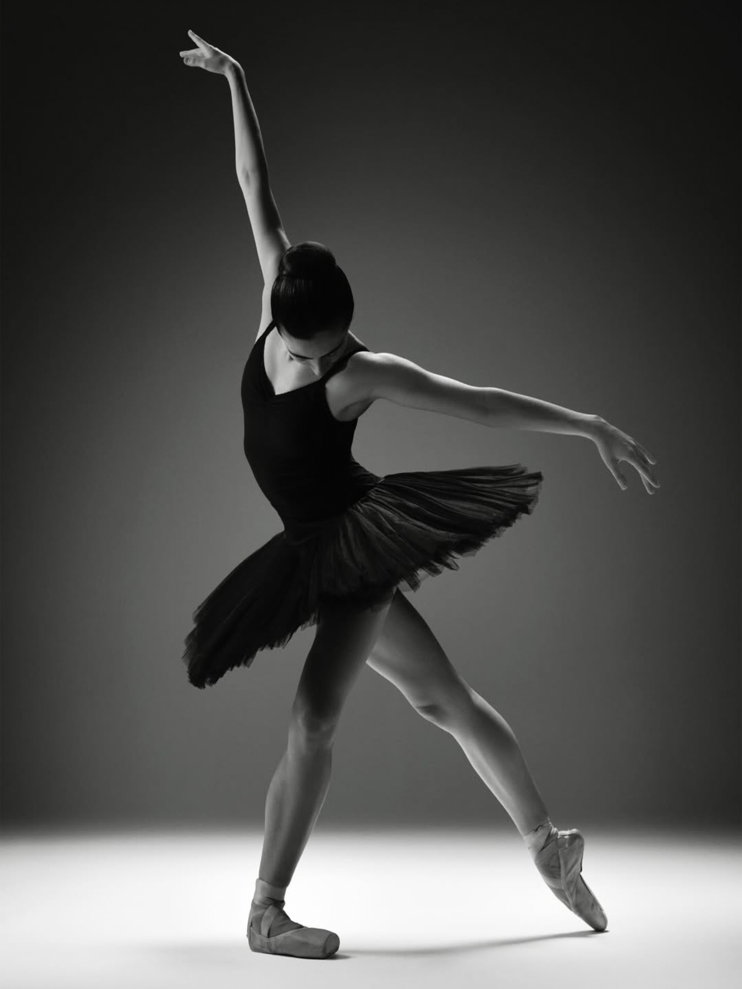 Black and white photo of a ballet dancer in a pose with one leg bent and raised, arms extended, wearing a tutu and pointe shoes.