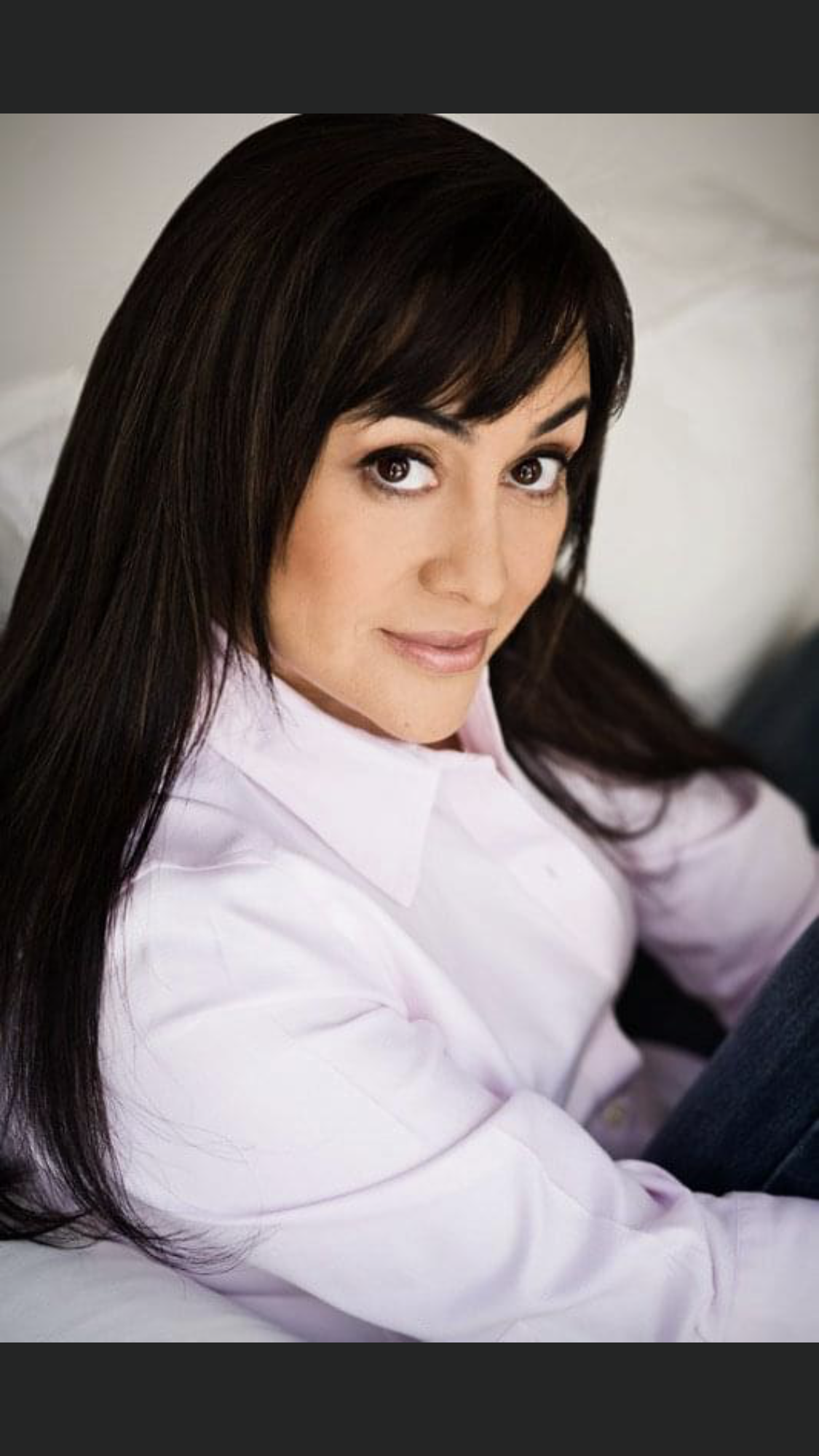 A woman with long dark hair and brown eyes wearing a light pink blouse, sitting on a sofa.