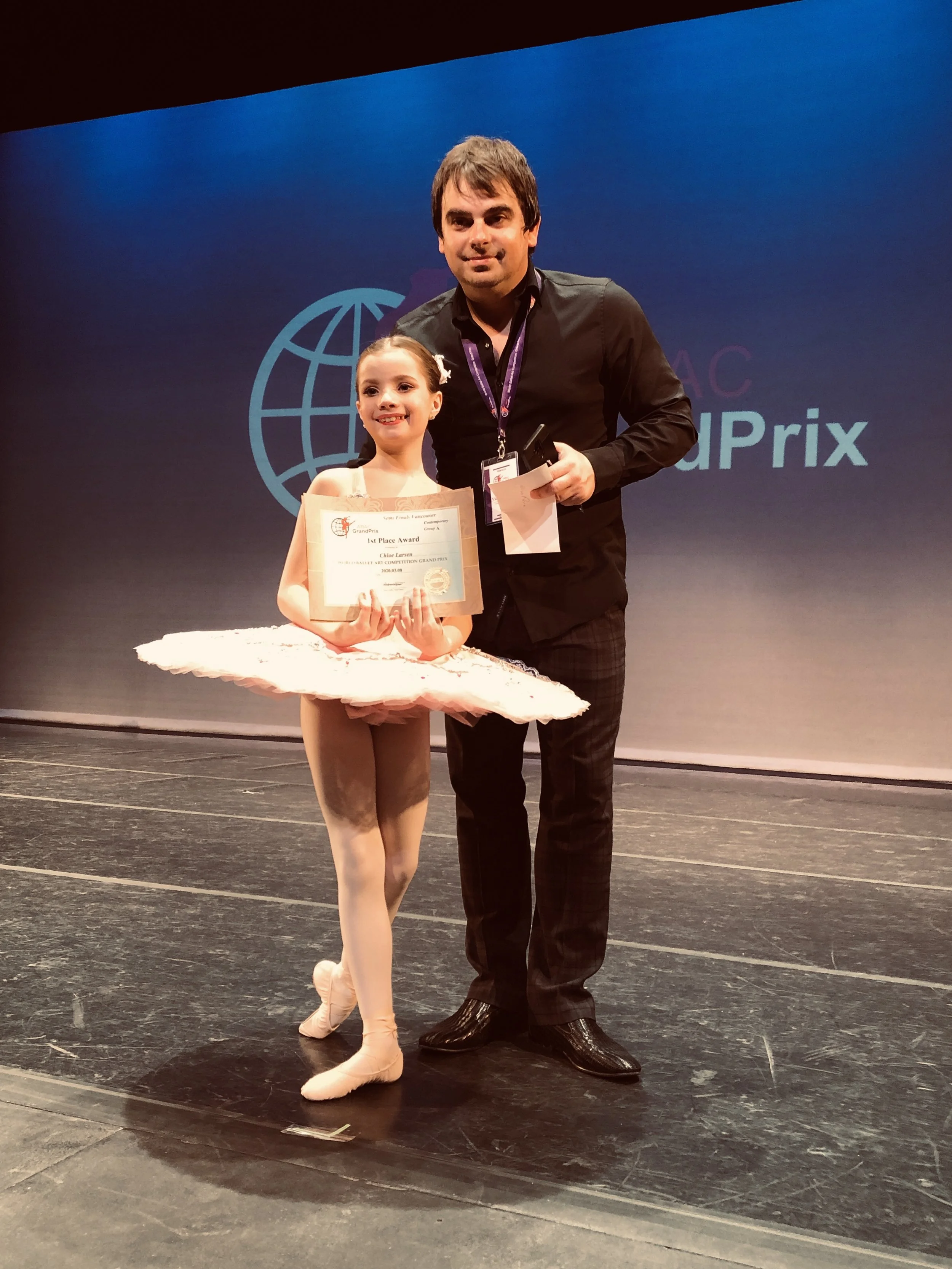 A young girl in a ballet costume holding a certificate, standing next to an adult man in a black shirt on a stage. The background displays a large logo with a globe and the words 'Grand Prix.'