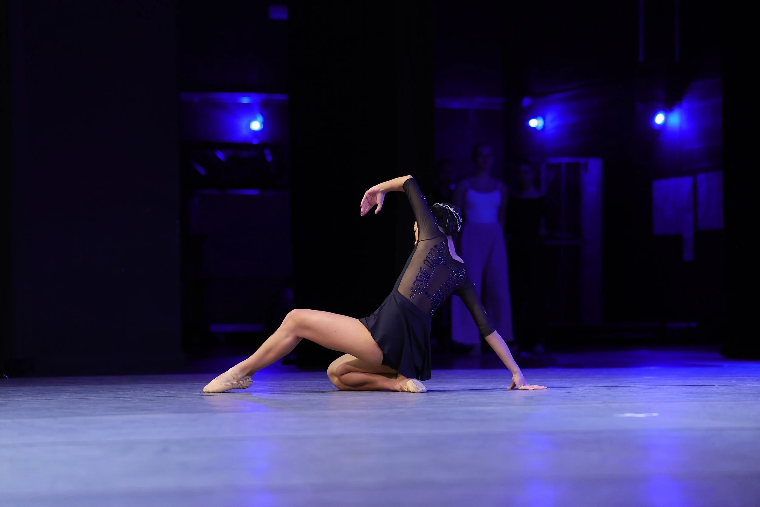 A ballet dancer in a black costume performing a pose on stage with a dark background illuminated by blue lights.