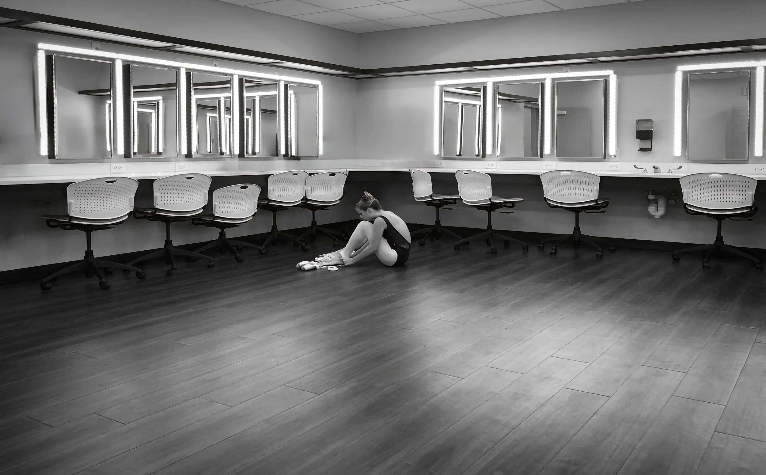 A girl in ballet attire sitting on the floor of a room with a long row of mirrors and chairs in the background.