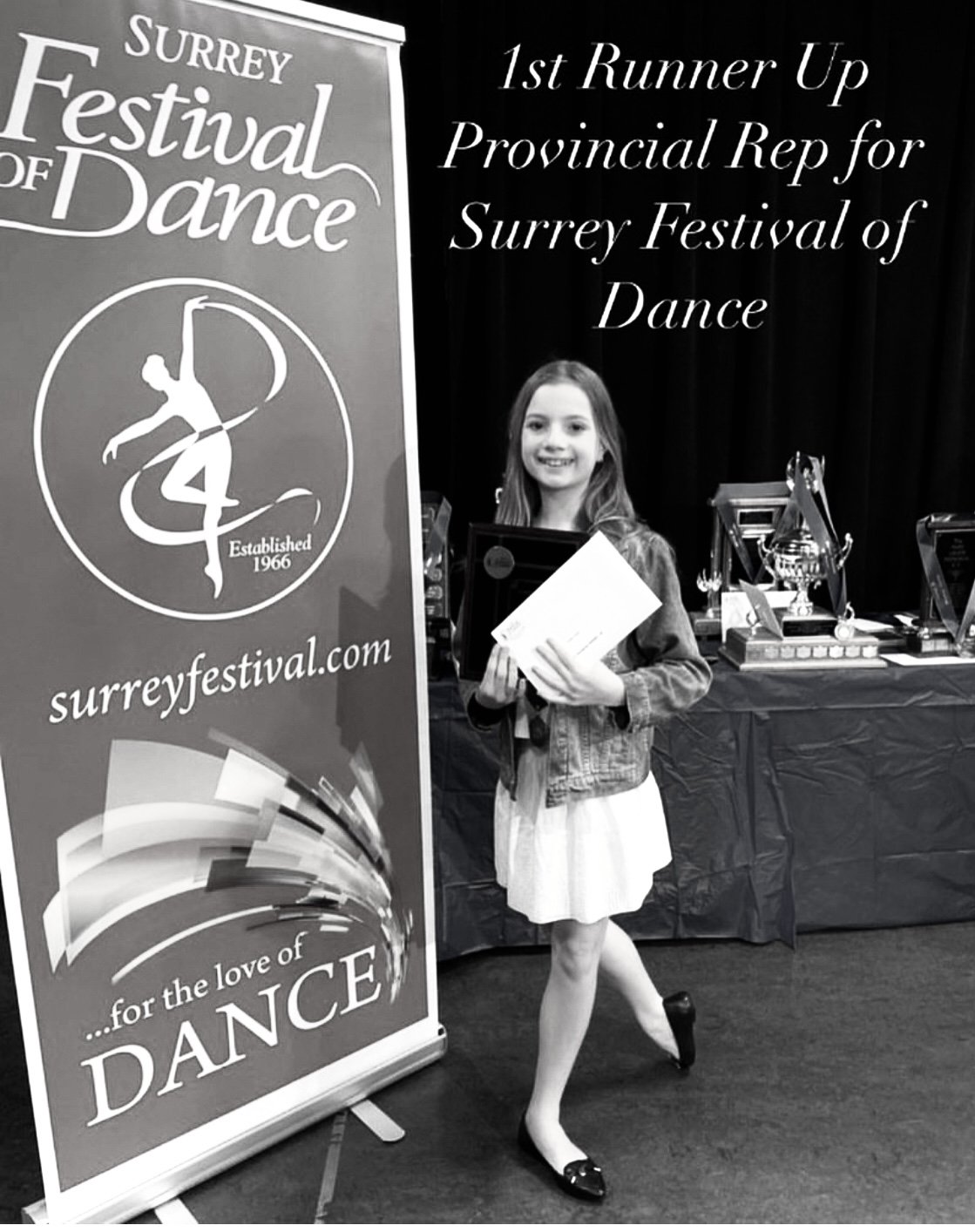 A young girl smiling and holding certificates, standing next to a banner for the Surrey Festival of Dance, with trophies on a table in the background.