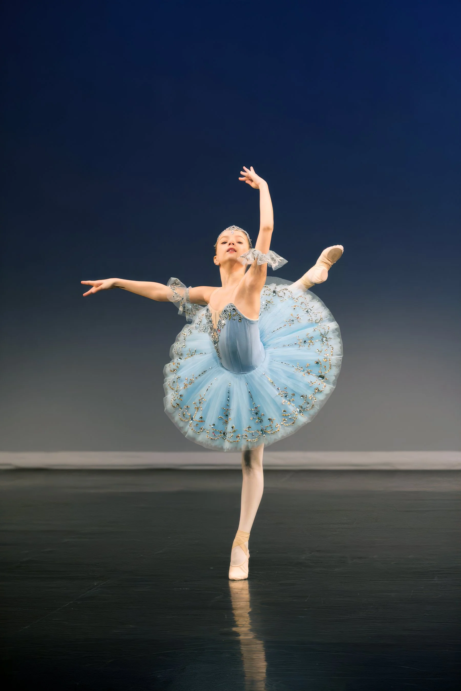 A young ballet dancer in a light blue tutu performing a ballet pose on stage with a dark background.