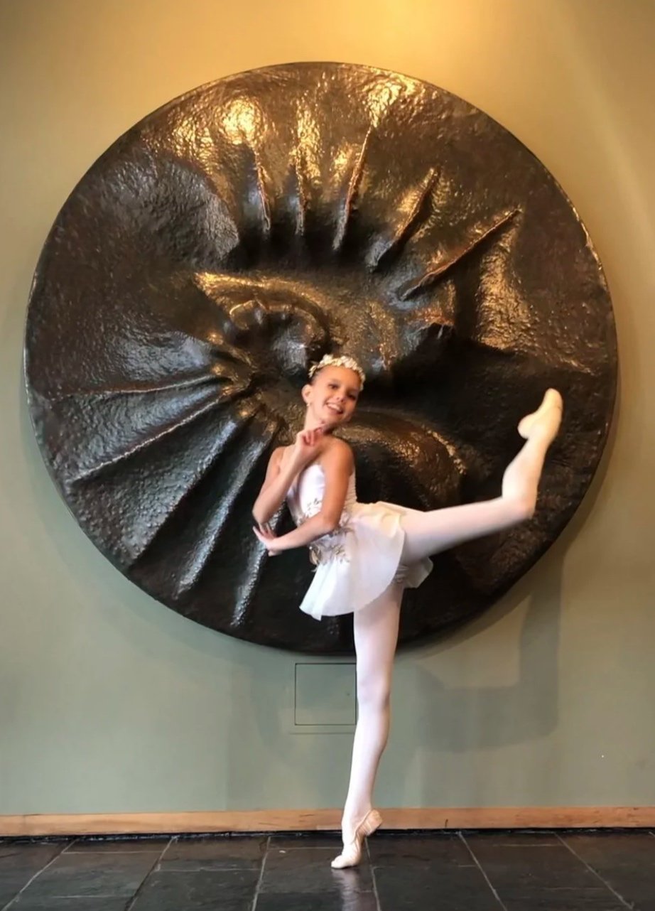 Young girl in ballet costume posing in front of a large, circular art sculpture with a spiral design.