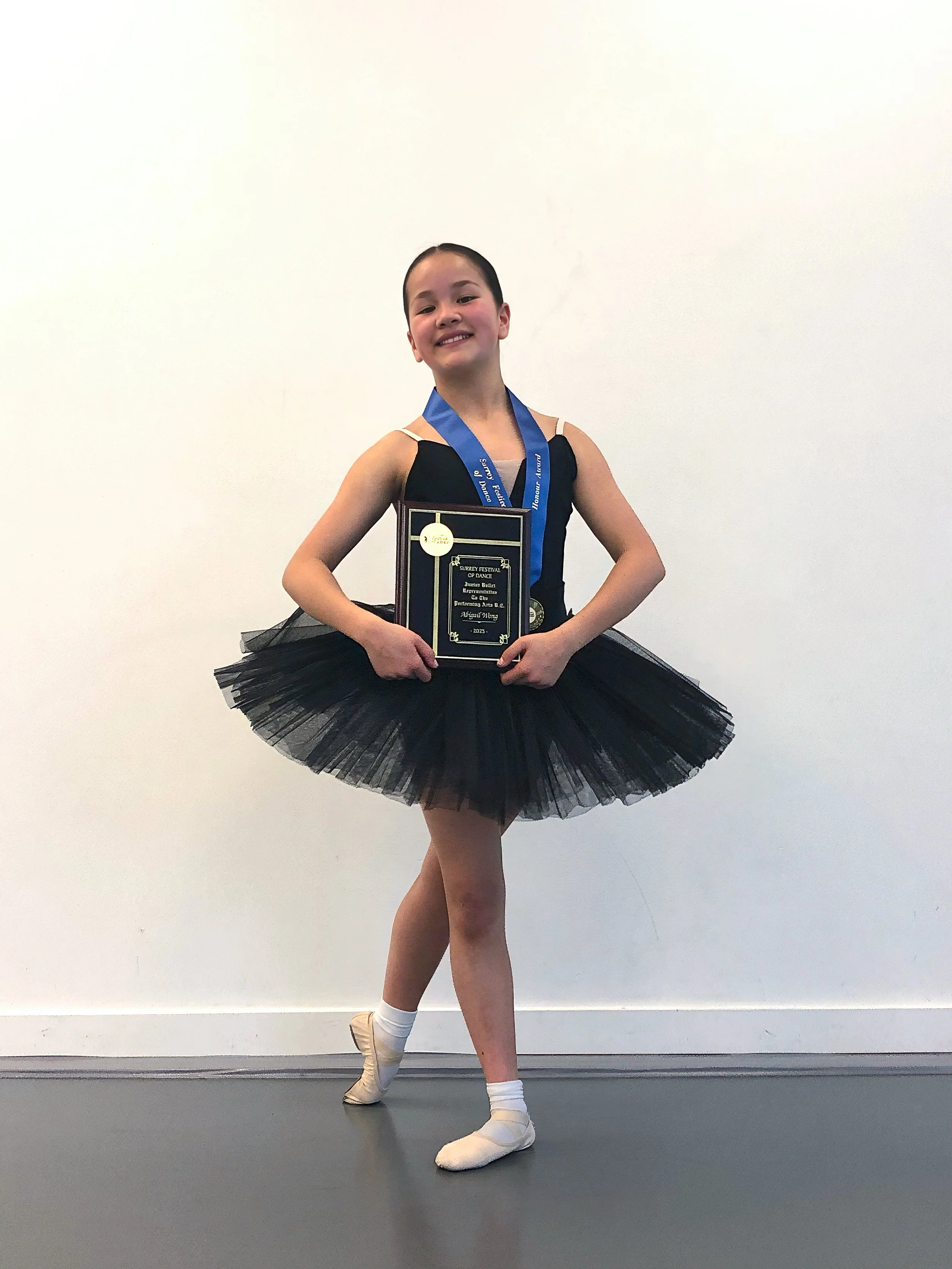 Young female ballet dancer in a black tutu and beige ballet slippers, standing on one leg in a ballet pose, holding a plaque and wearing a medal, against a plain white wall.