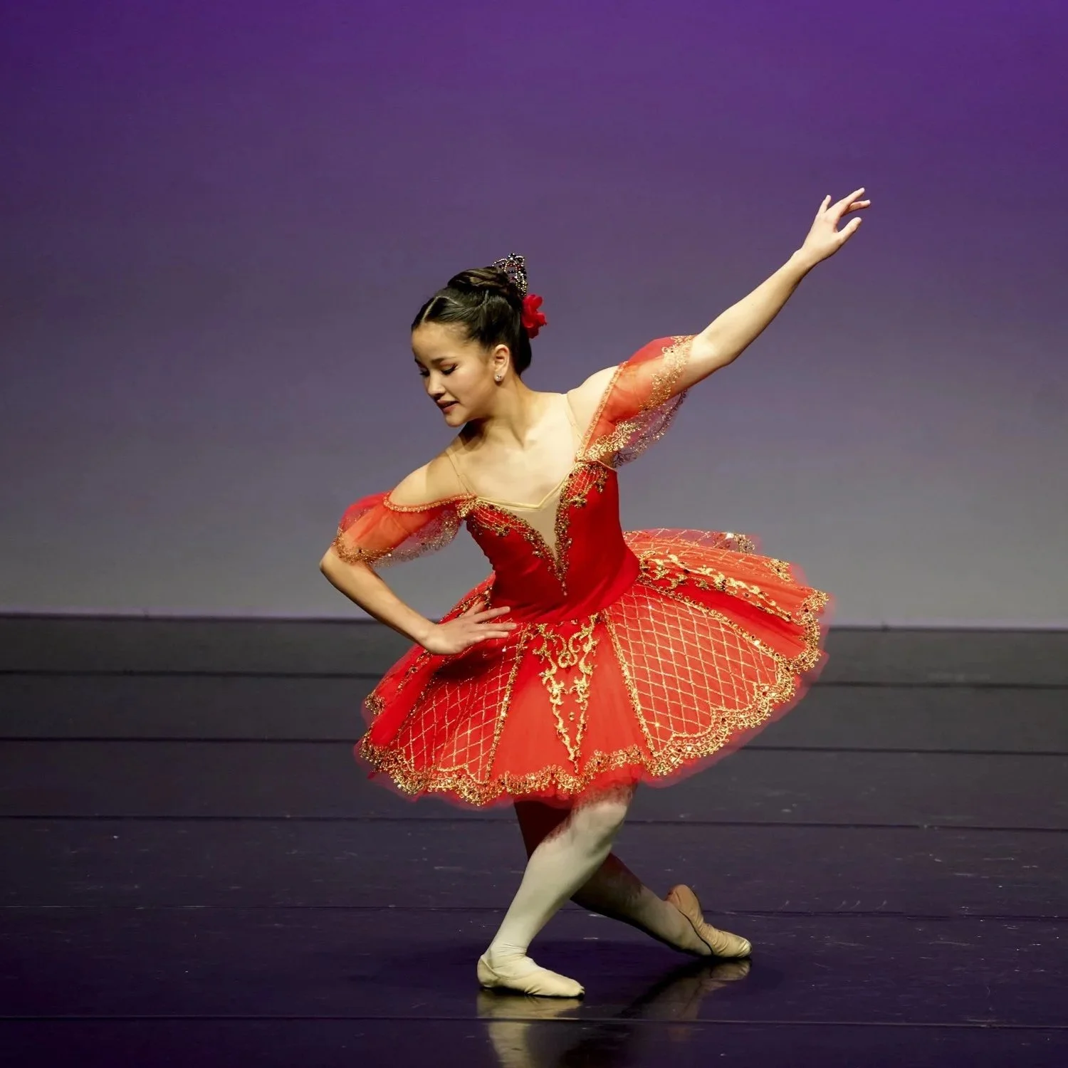 A ballerina performing on stage, dressed in a red tutu with gold embroidery, against a purple background.