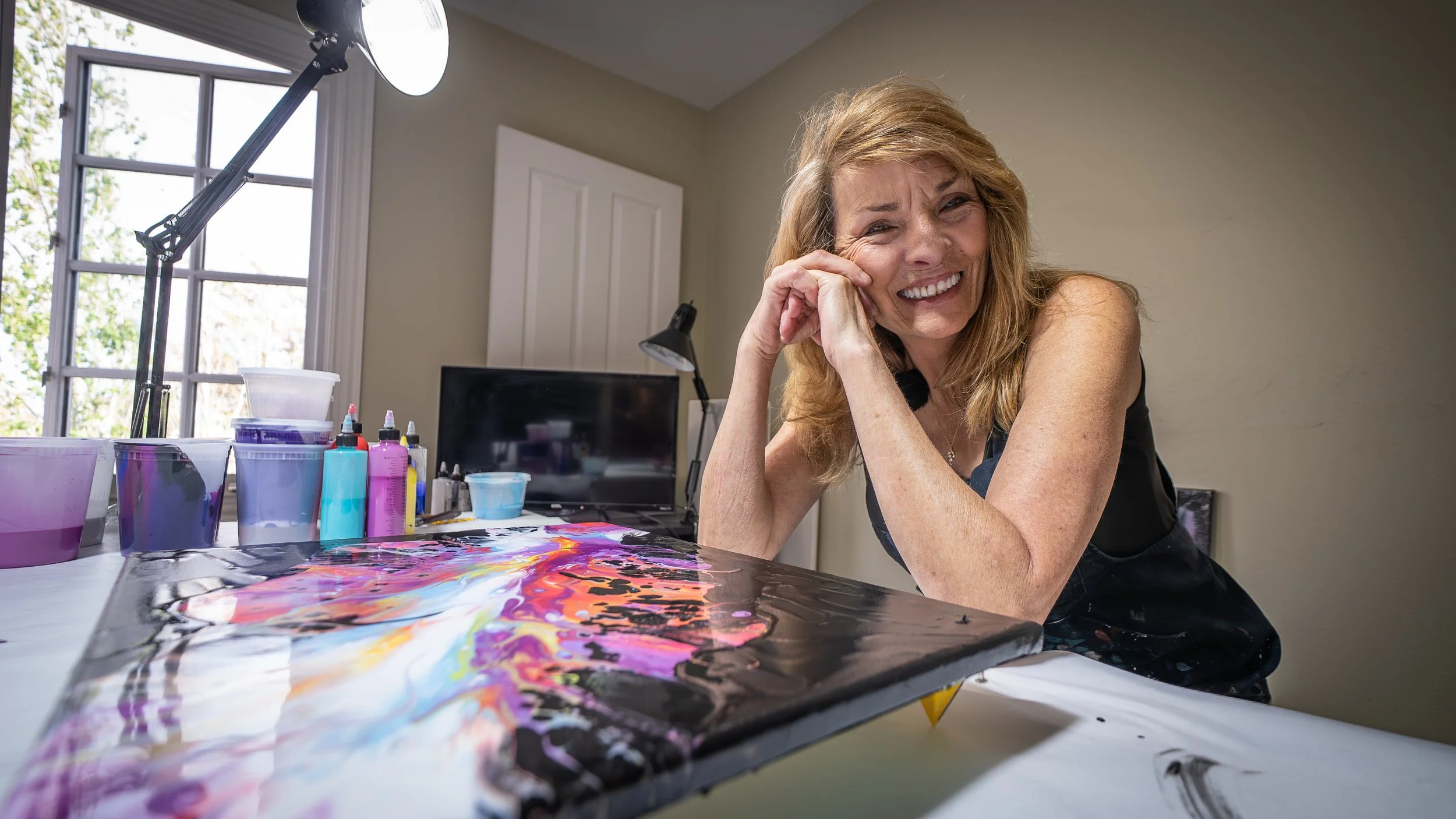 A woman with red hair smiling and resting her head on her hand at a table with colorful paint supplies and a canvas.