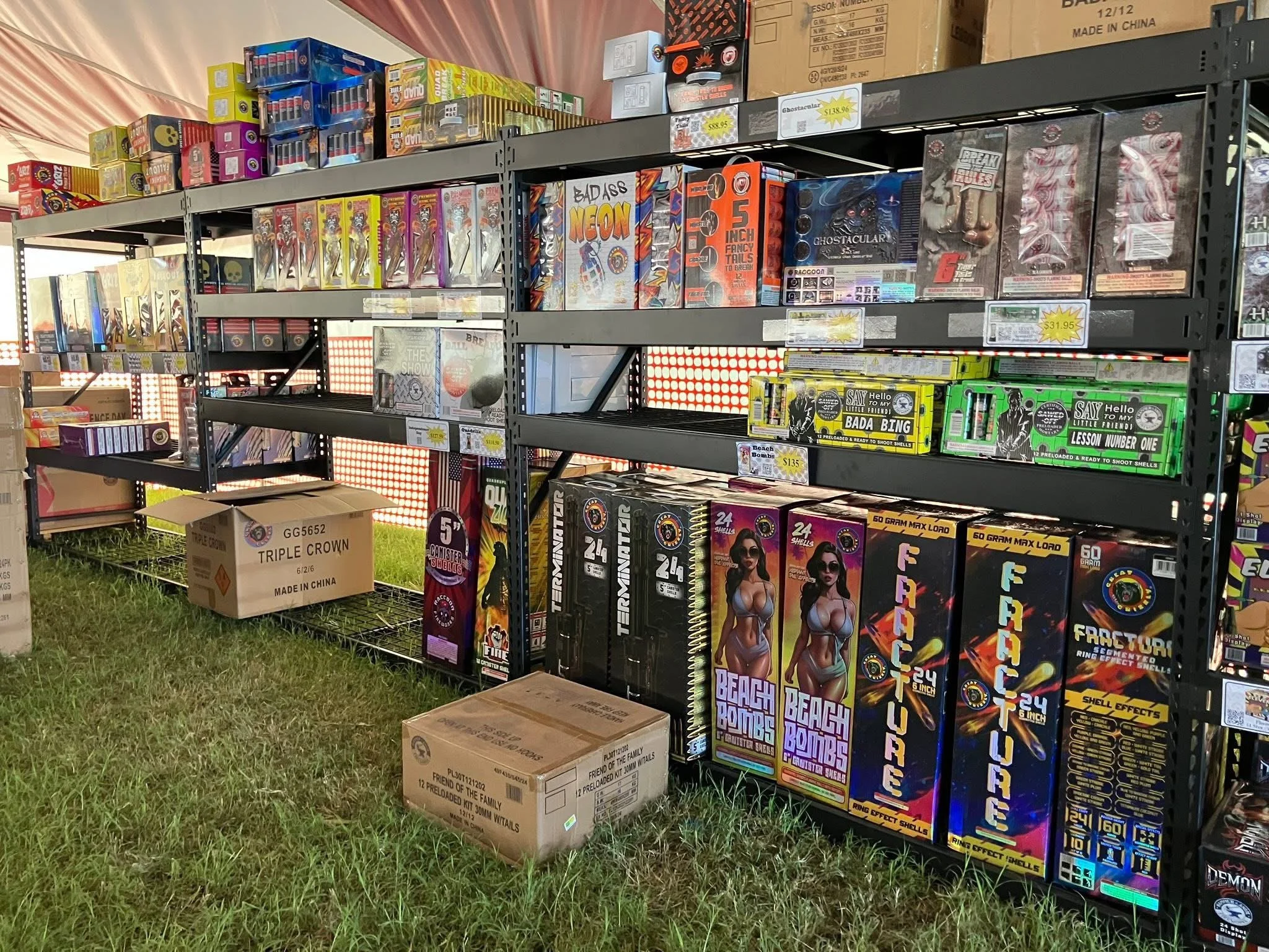Shelves stocked with fireworks, including boxed fireworks labeled 'Beach Bombs' and 'Shell Effects', under a tent with grass underneath. Several cardboard boxes sit on the ground in front of the shelves.