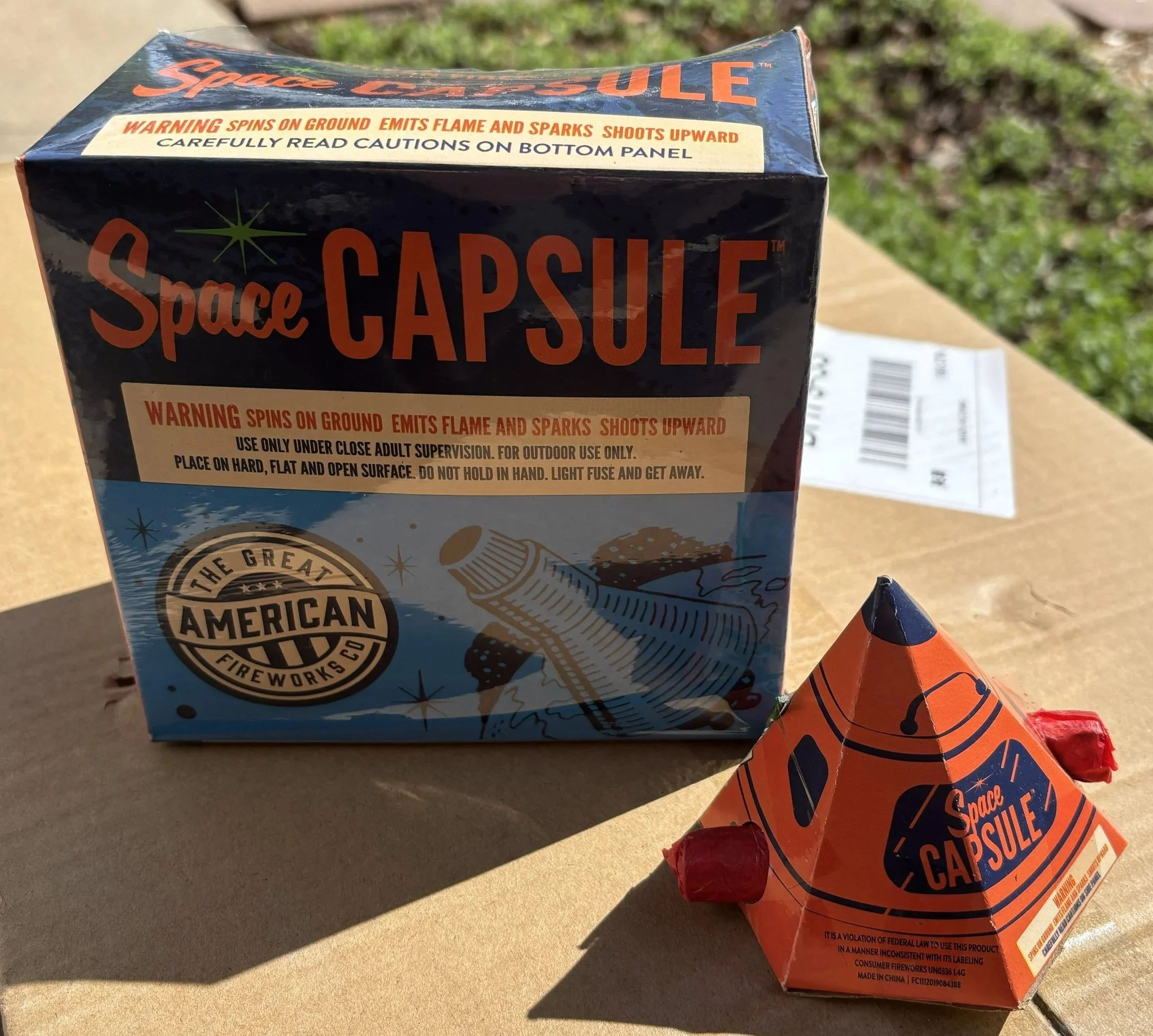 Boxes of Space Caution fireworks with warning labels, placed outdoors on a concrete surface with grass in the background.