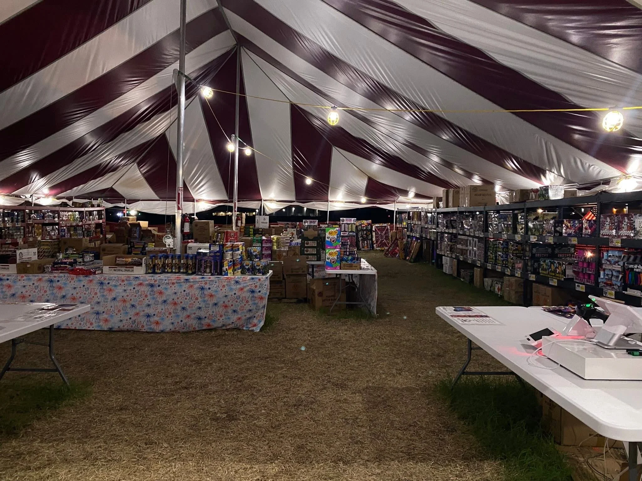 Nighttime view of a large event tent with maroon and white stripes, lit by hanging string lights. Beneath the tent, there are tables and shelves filled with various boxes and fireworks for sale or display, including a table with patriotic-themed decoration.