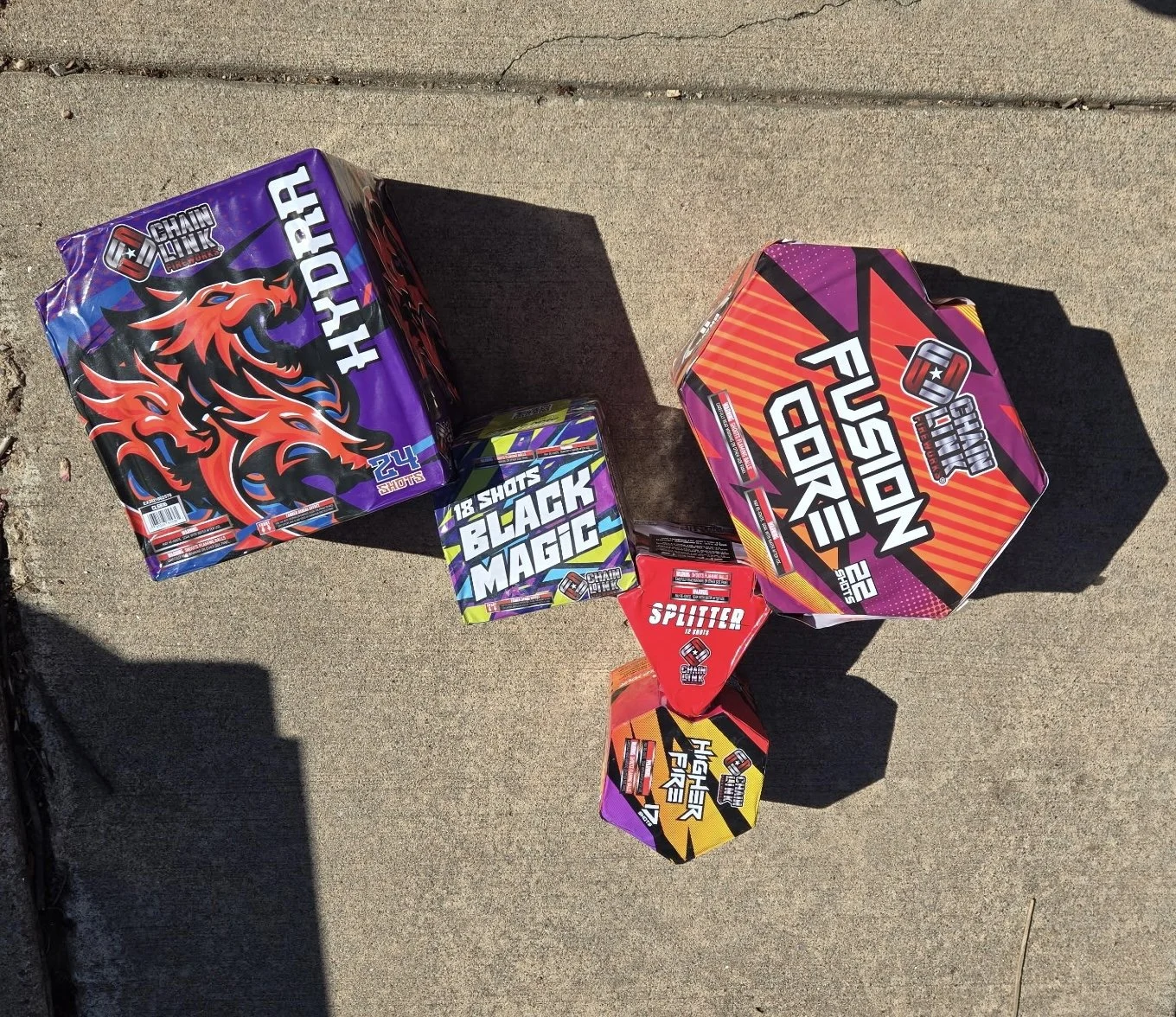 Several fireworks boxes with colorful packaging, including one labeled 'HYPER FIRE,' another 'BLACK MAGIC,' one 'POP CORN,' one 'SPITTER,' and one 'HIGHER FIRE,' placed on a concrete sidewalk.