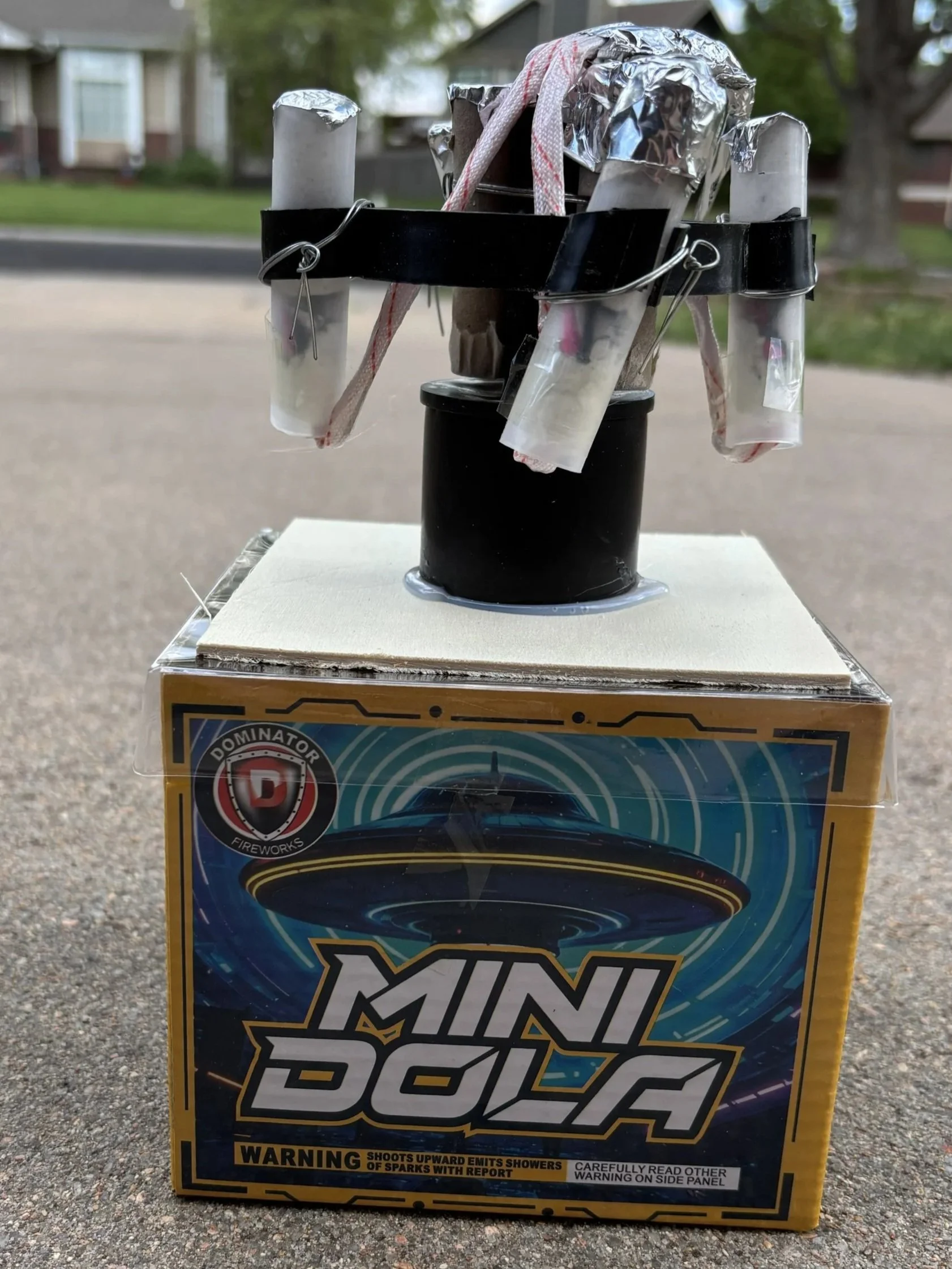 Firework box labeled 'Mini Dola' with a small black device on top, several tubes, and attached fuses.