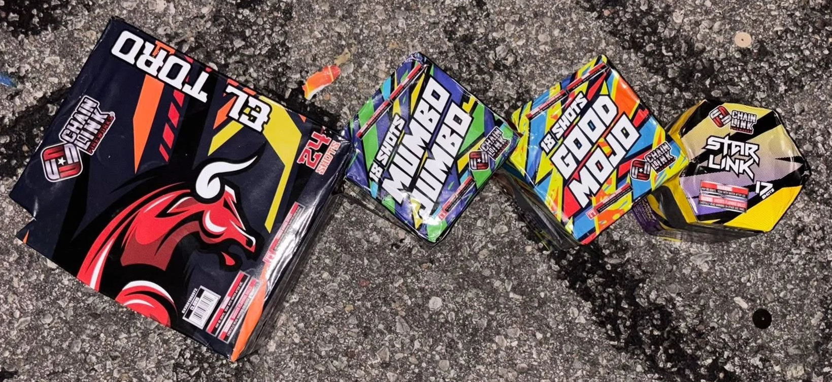 Four brightly colored packs of fireworks with various designs and labels, laying on an asphalt surface.