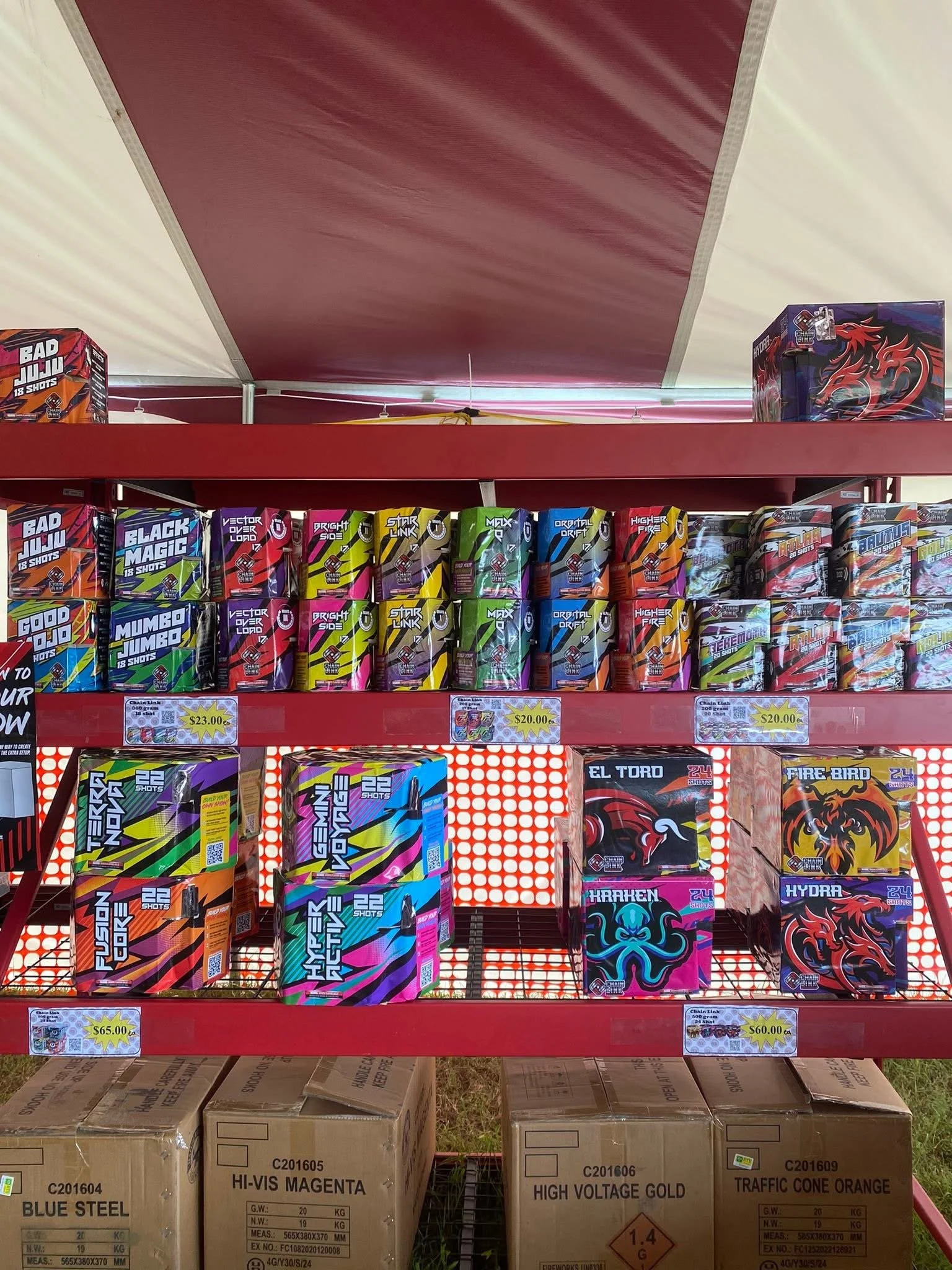 Shelves of fireworks boxes in a store, with colorful labels including 'Bad Juju,' 'Black Magic,' 'Star Link,' 'Hydra,' and 'Krakken.' The boxes are on red metal shelves, with some larger boxes on the bottom shelf. Price tags indicate cost per box.
